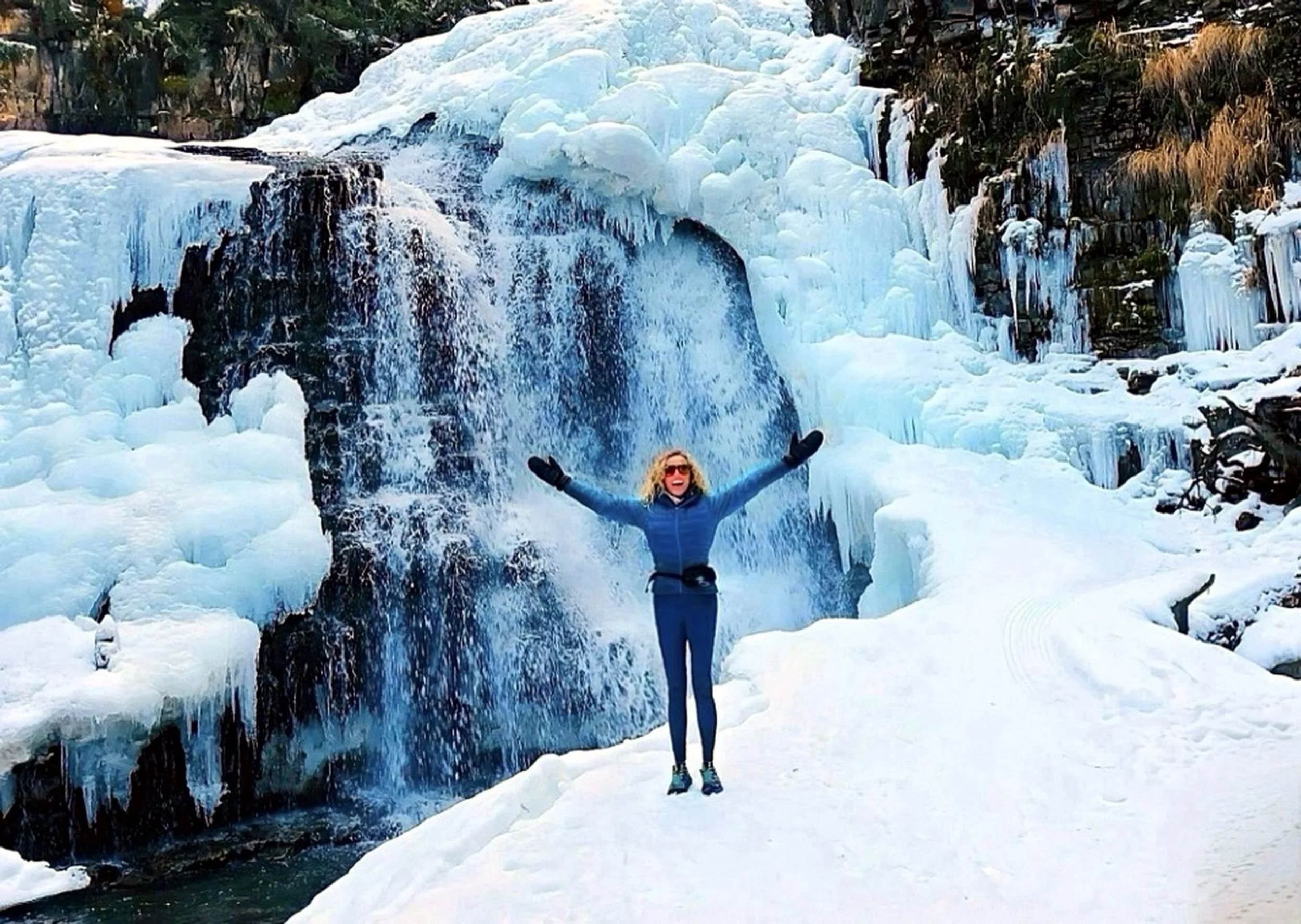 A woman wearing a blue winter jacket and black gloves stands with her arms outstretched in front of a partially frozen waterfall in a snowy landscape.