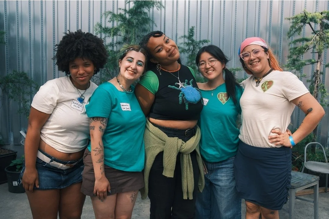 Five people standing together outdoors in front of a metal wall and trees, smiling at the camera.