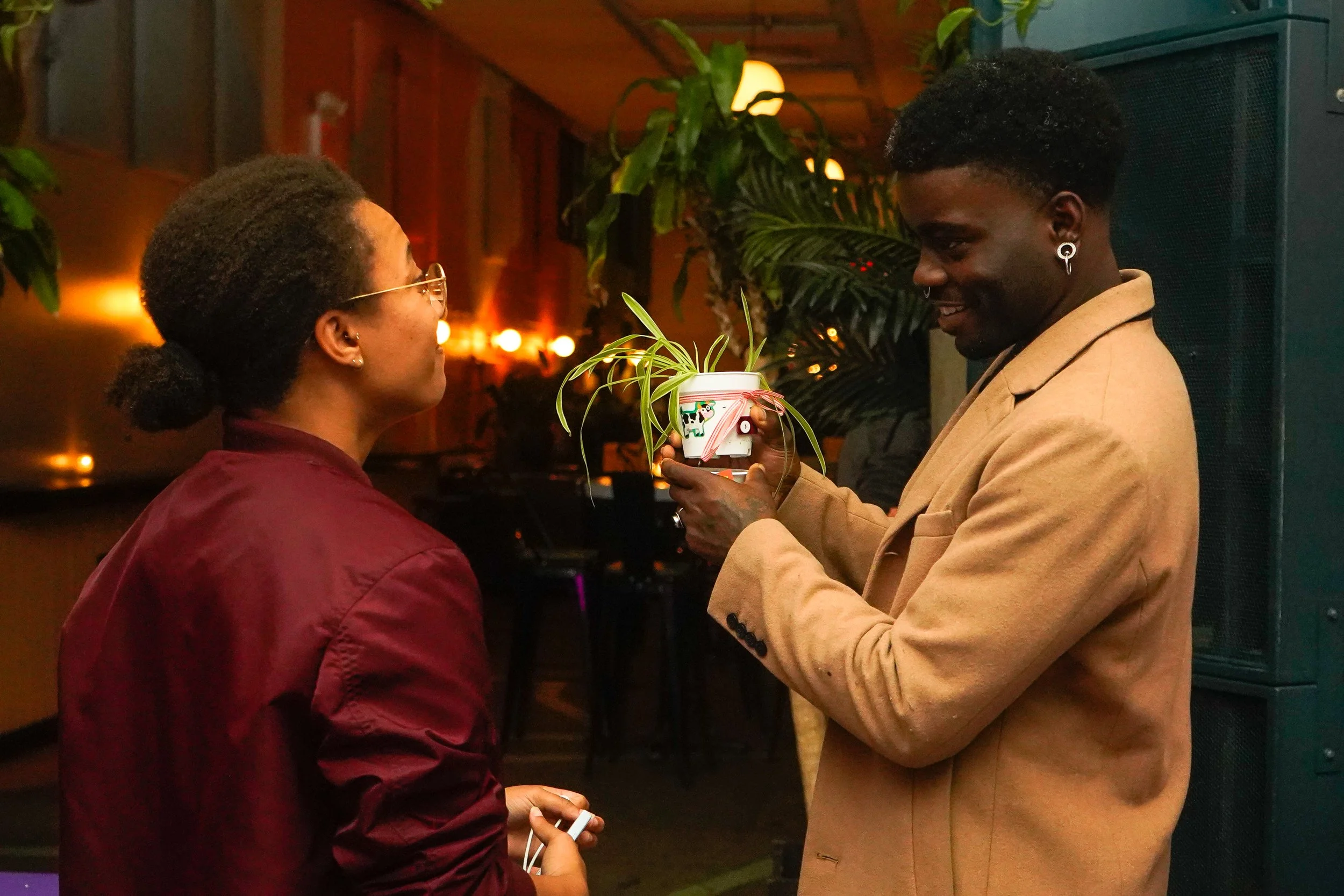 A man is giving a woman a potted plant as a gift at an indoor event, with warm lighting and greenery in the background.
