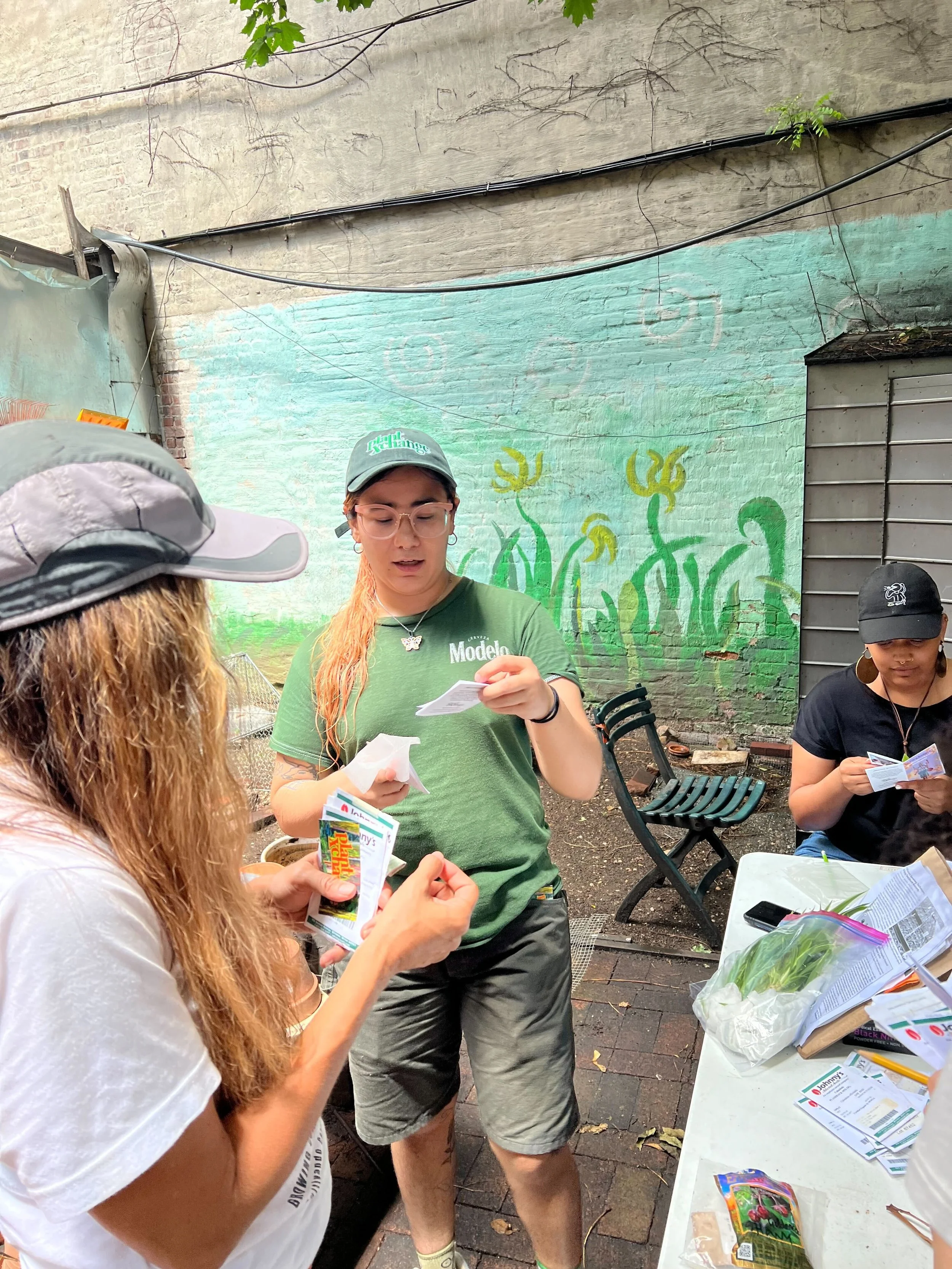 A woman with short blonde hair, wearing an orange sleeveless top and beige pants, sitting on a bench and writing in a notebook. There is a large potted plant and abstract art on the wall behind her, with sunlight casting shadows.