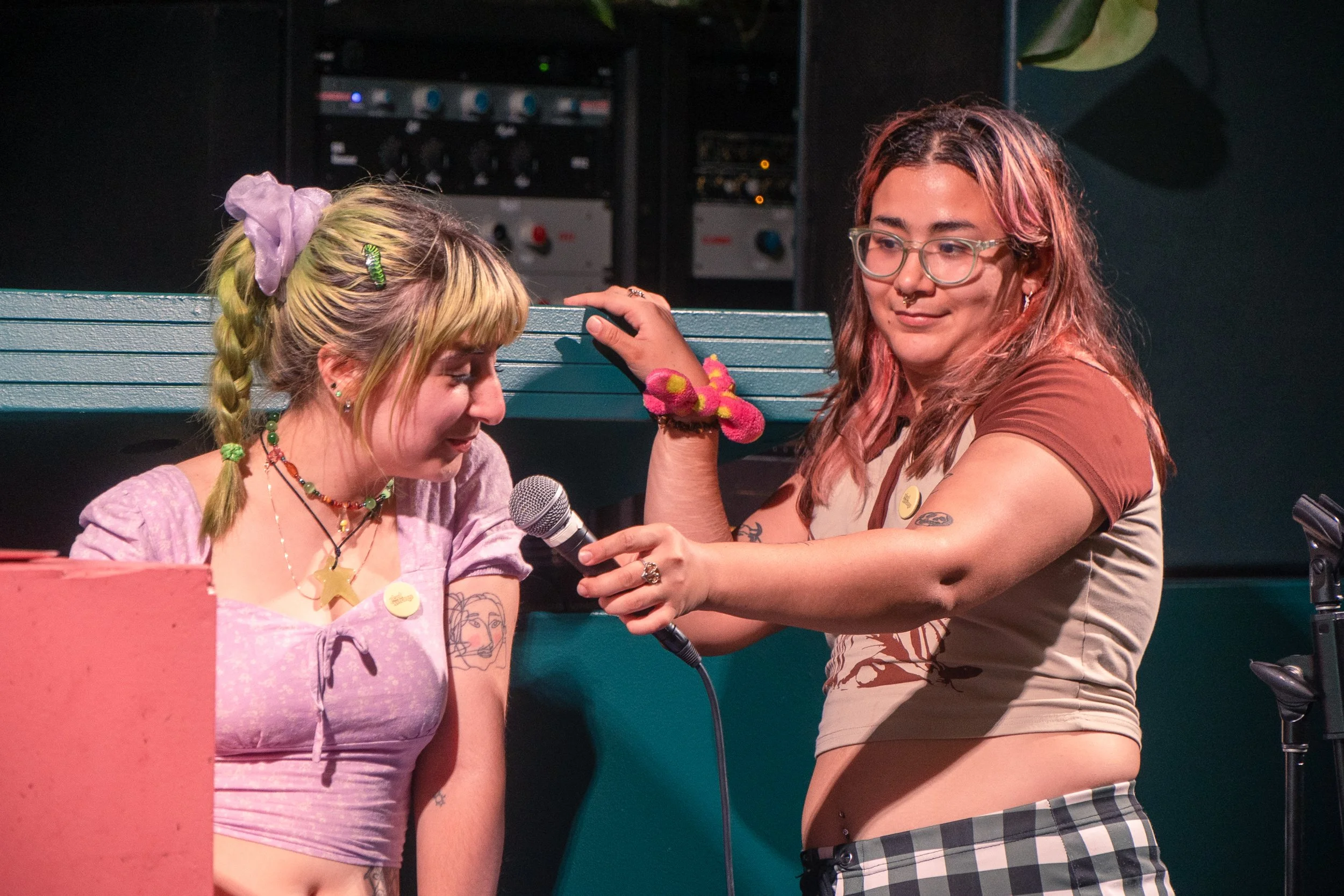 Two young people on stage, one with blonde hair and pastel purple clothing, the other with reddish hair and glasses, engaging in an activity involving a microphone and a casual pose, with a DJ setup in the background.