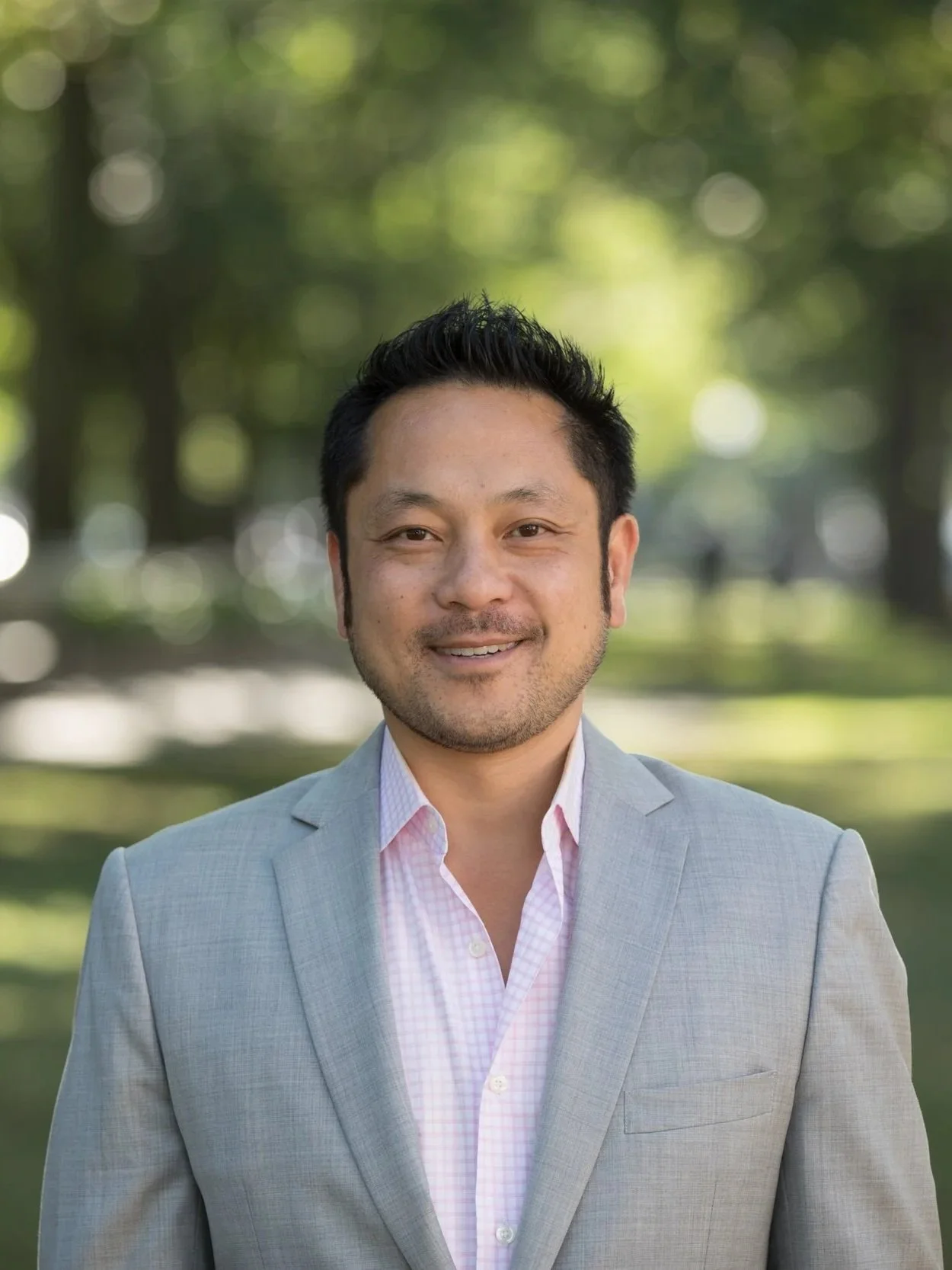 Portrait of a man wearing a light gray blazer and pink checkered shirt, standing outdoors with blurred trees and greenery in the background.