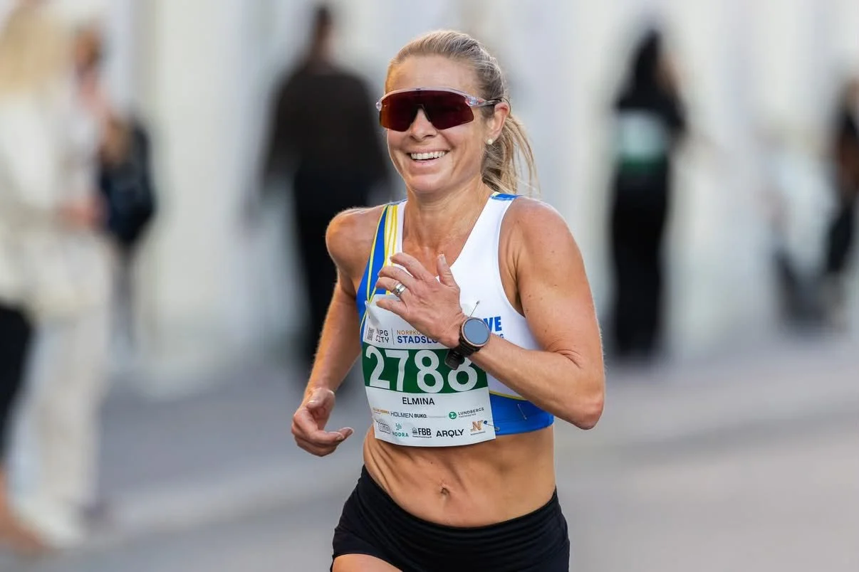 A woman running in a race, wearing a white sports top with blue accents, black shorts, sunglasses, and a smartwatch, smiling as she nears the finish line.