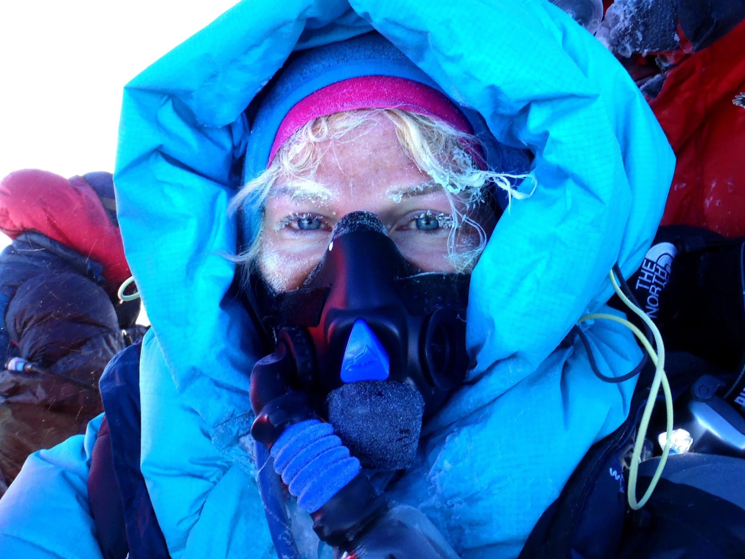 Close-up of a person's face wearing a blue hood and ski goggles, with snow on their face and blonde hair, in a snowy outdoor setting.