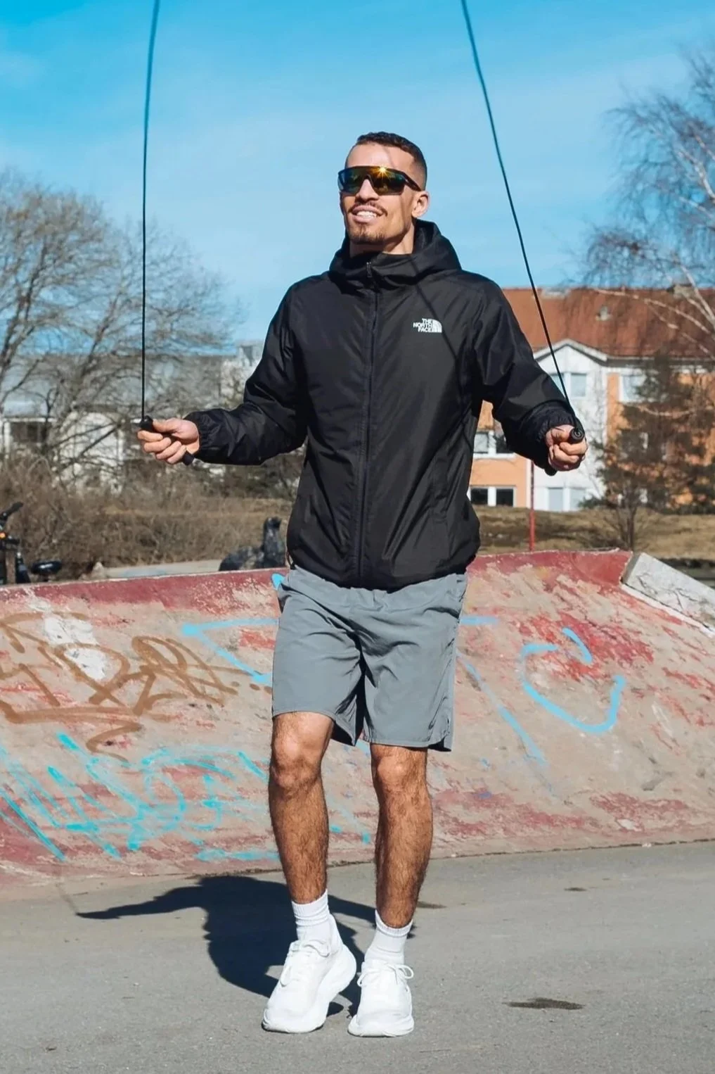 A man skipping rope at a skate park during daytime, wearing sunglasses, a black jacket, gray shorts, white sneakers, and white socks.