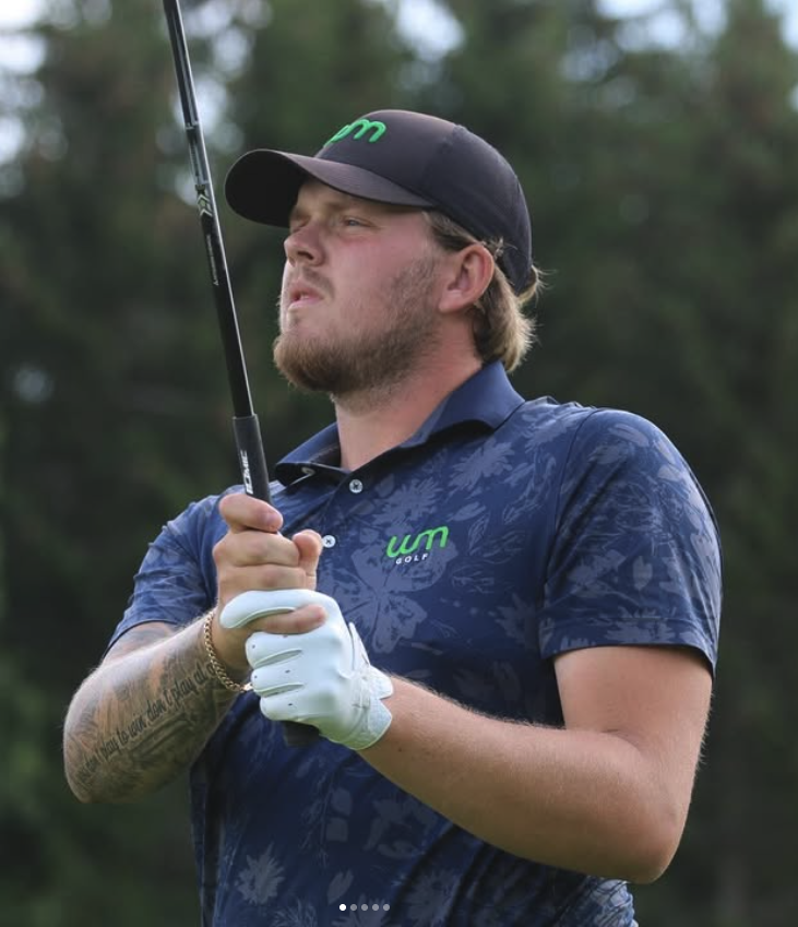 A man in a blue golf shirt and black cap holding a golf club on a golf course, with trees in the background.