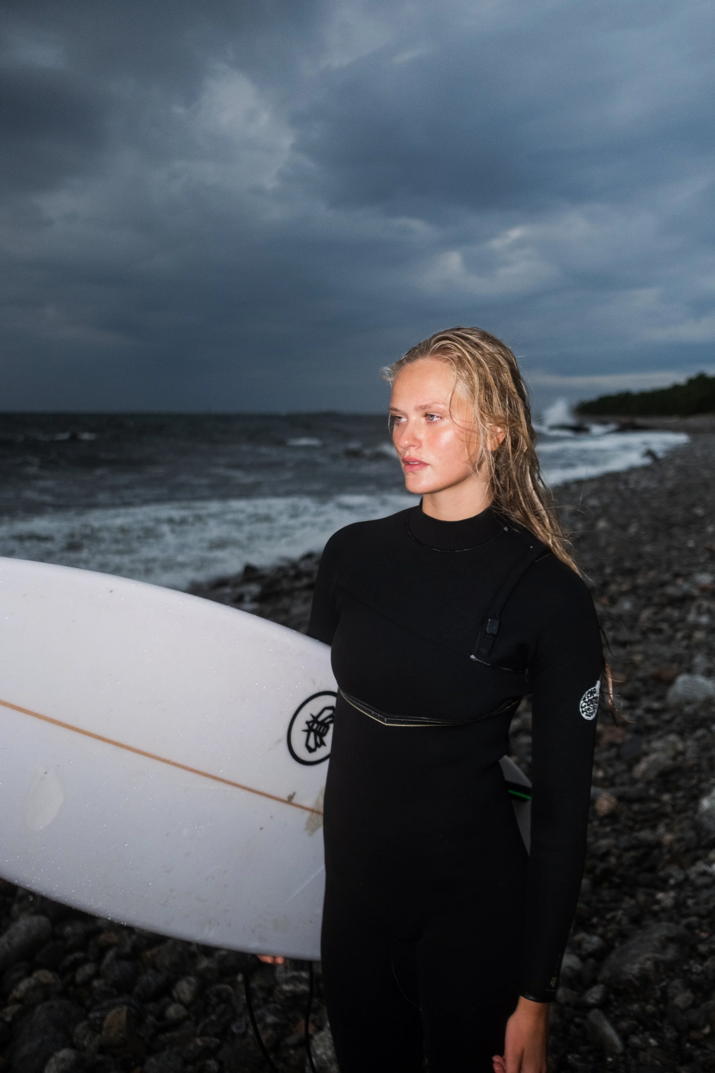A young woman with blonde, wet hair wearing a black wetsuit standing on a rocky beach holding a white surfboard, with dark cloudy skies and the ocean in the background.