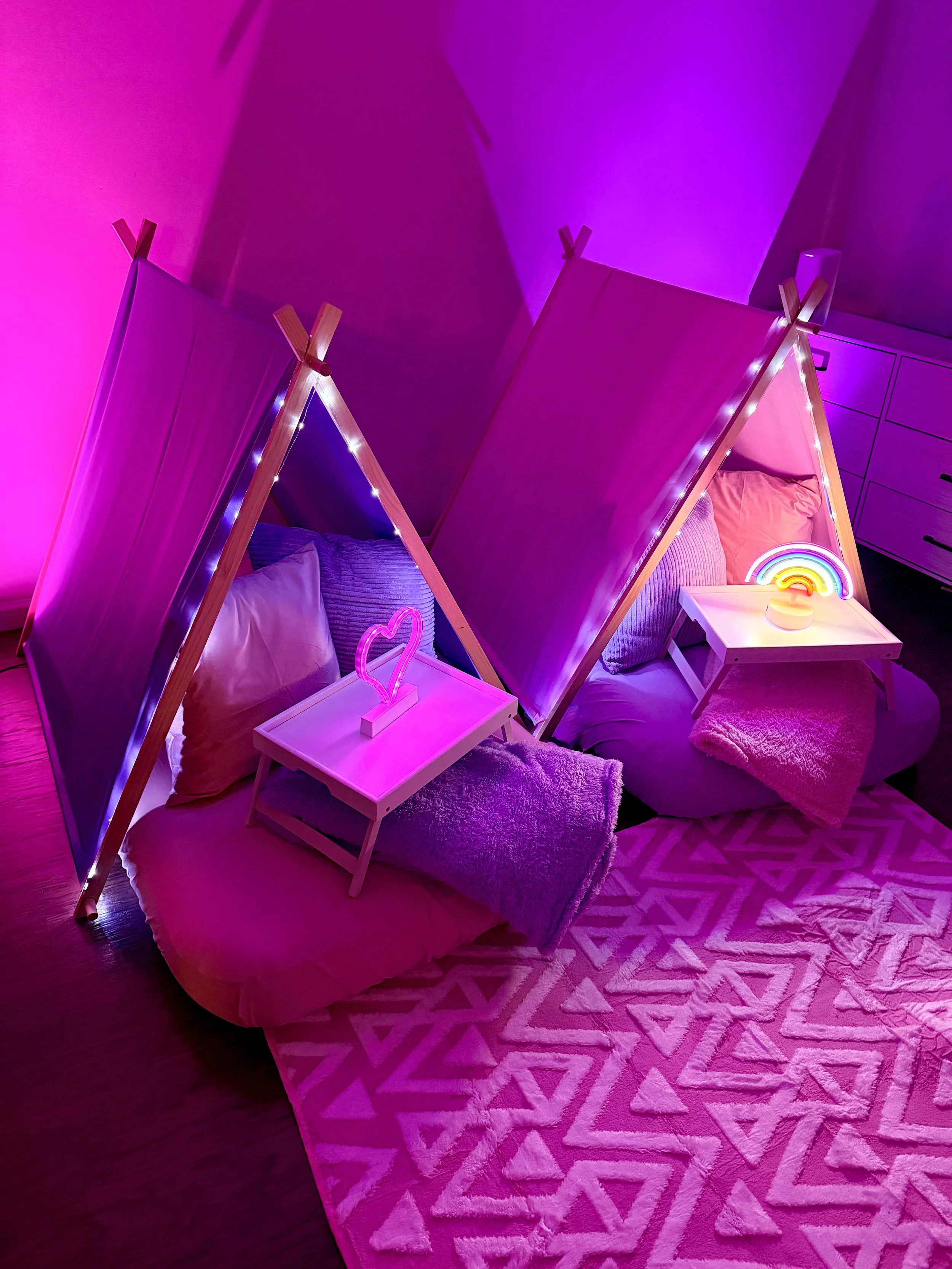 Two beds with wooden frames and small white bedside tables decorated with neon lights in the shape of a heart and a rainbow, in a room with purple and pink lighting.