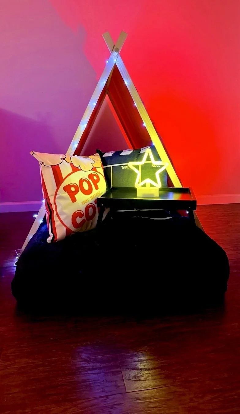 A cozy corner with a small tent made from wooden sticks, illuminated with string lights. Inside, there are popcorn-themed and striped pillows, a black side table with a neon star sign, and a dark bean bag on a wooden floor, with colorful wall lighting.