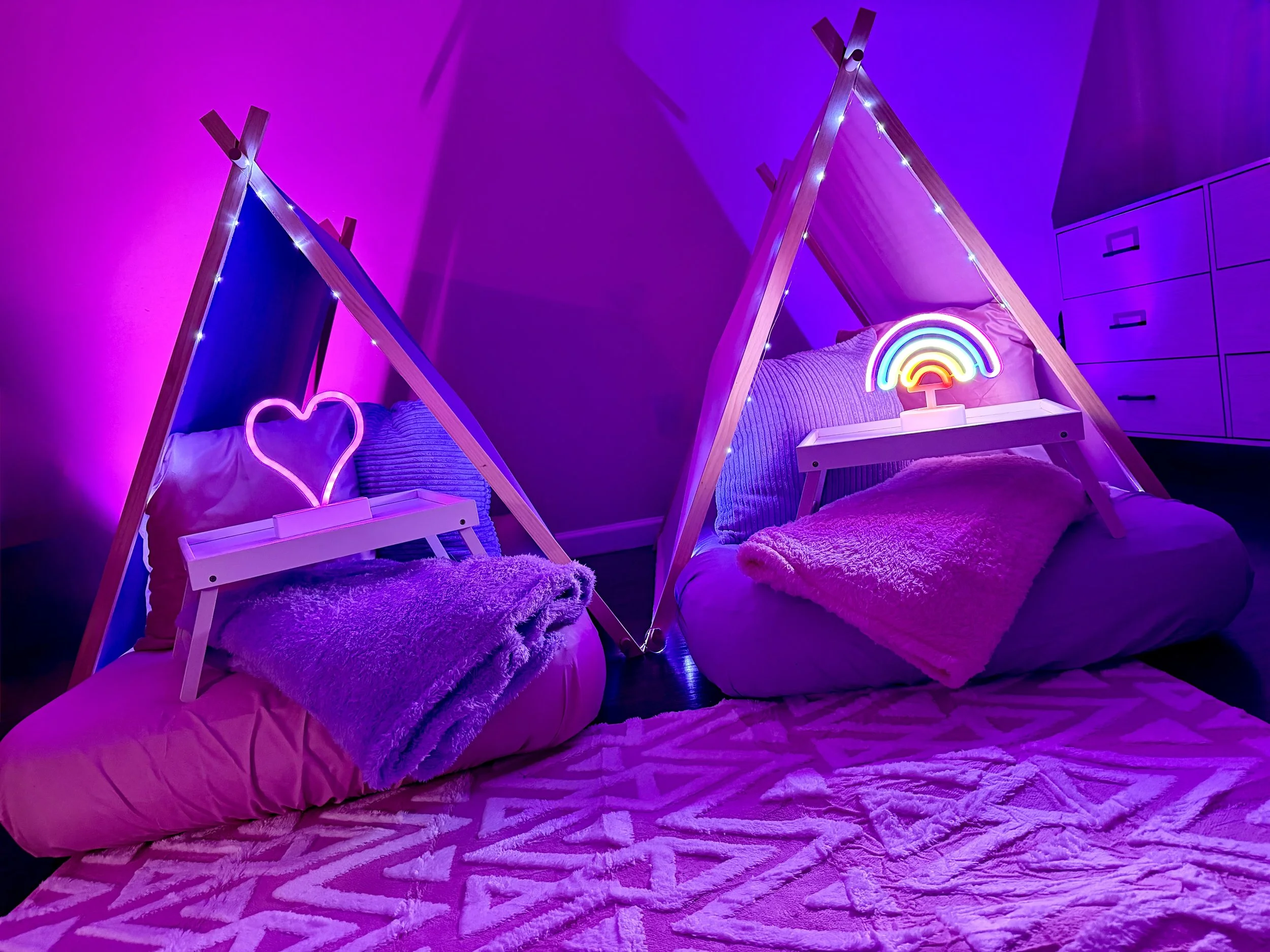 A cozy bedroom scene with two bed setups, each with a small wooden tent-like structure illuminated by colorful neon lights, a heart on one side and a rainbow on the other. The bedding is pink and purple, with plush blankets and striped pillows. The room is bathed in purple and pink lighting.
