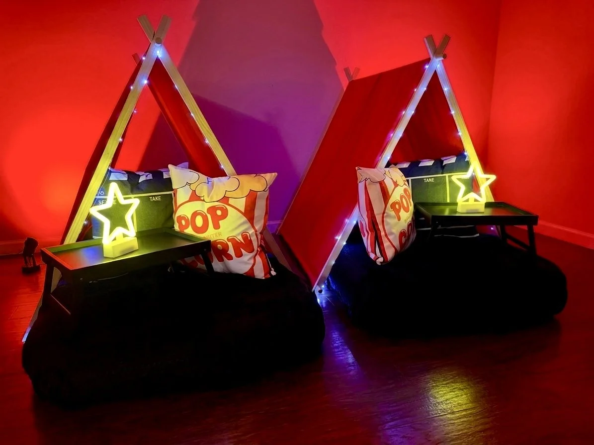 Two indoor seating areas designed as small tents with red sides, supported by wooden frames, lit with string lights, each with a black pillow, a star-shaped lamp, and a popcorn pillow, set against a red wall.