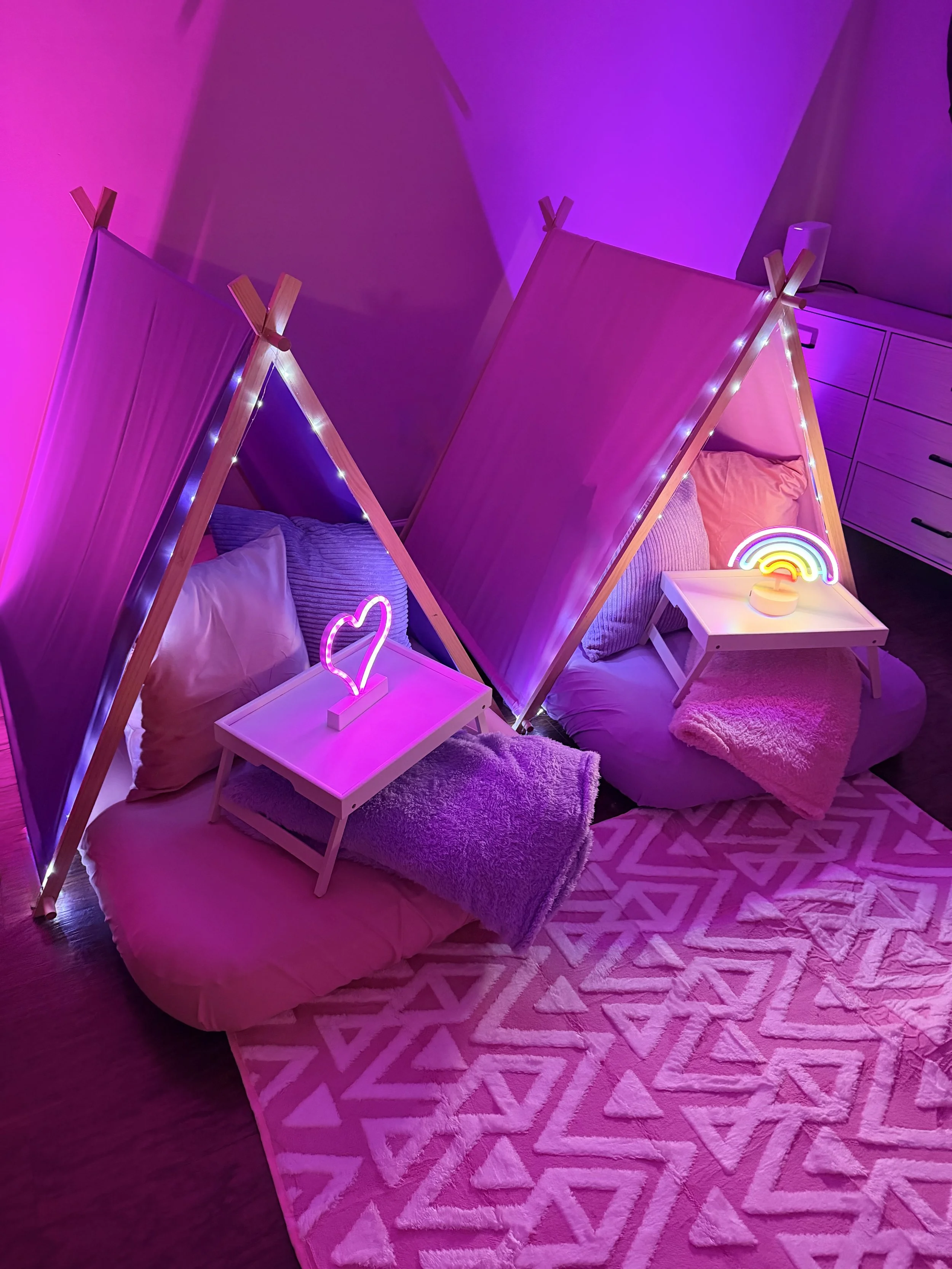 Two beds with triangular pink fabric privacy screens, each with pastel pillows, small white nightstands with neon heart and rainbow signs, soft pink and purple lighting, and a geometric pink rug on dark wooden floor.