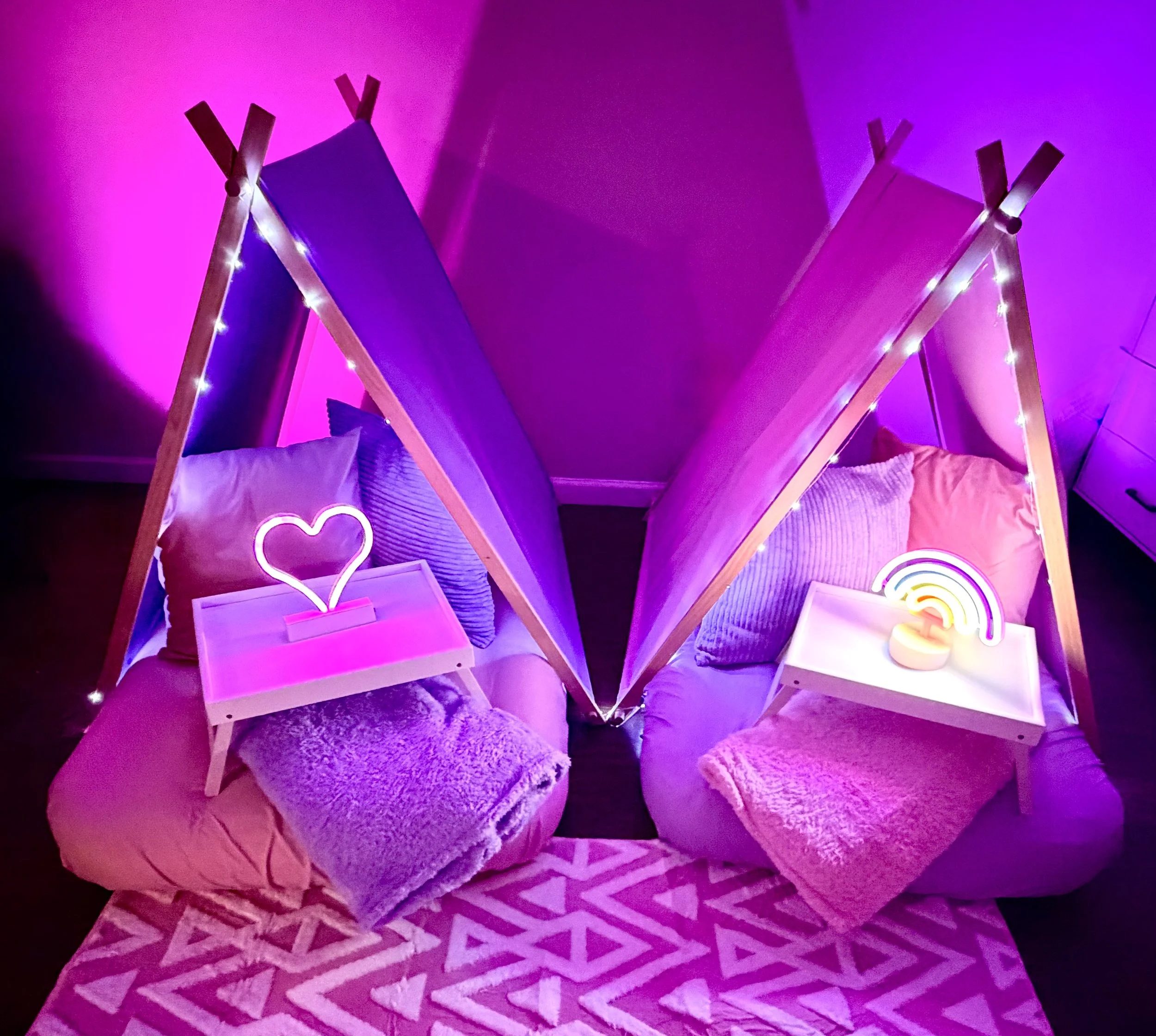 Two bean bag chairs with pillows and plush blankets, each with a small LED table featuring neon decor and string lights, set in a pink and purple-lit room.