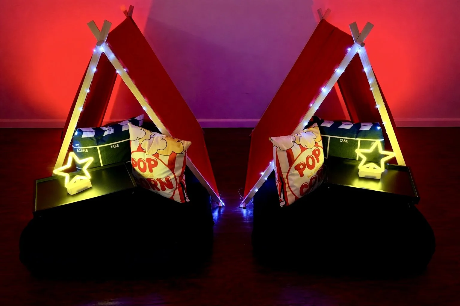 A cozy indoor scene with two small red tents lit up with string lights, each containing a pillow shaped like a popcorn bag, neon star-shaped lights, and a black surface underneath, against a wall lit with red and purple ambient lighting.