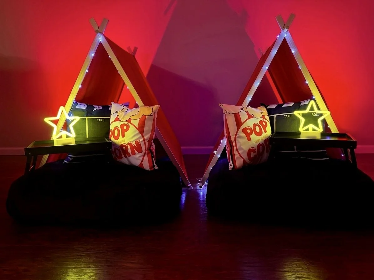 Two small indoor tents decorated with string lights, each with a neon star-shaped light and popcorn-themed pillow, placed on bean bags with small tables on side.