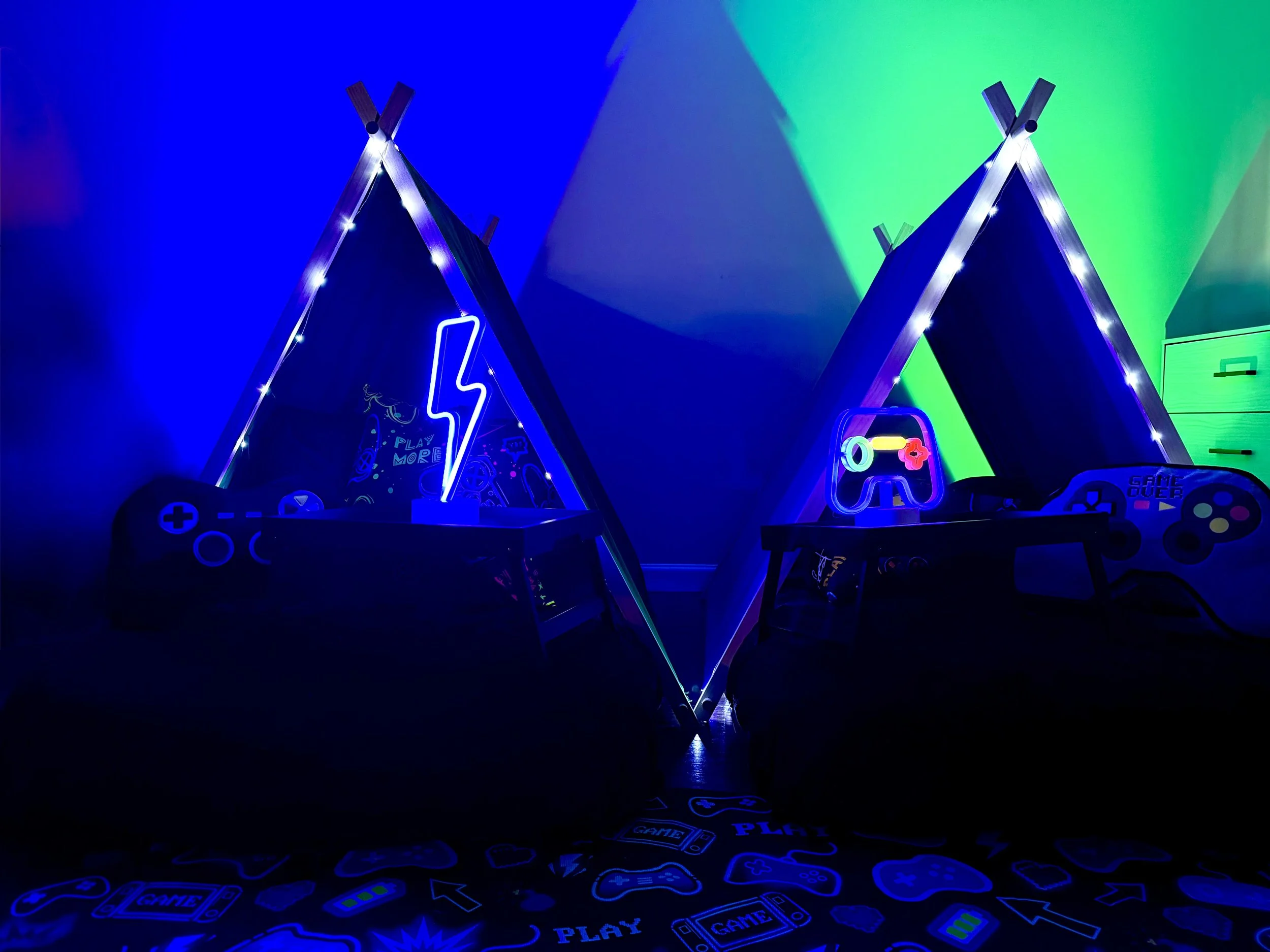 A gaming setup with two blue LED-lit tents resembling teepees, decorated with neon gaming-themed signs and controllers, illuminated with blue and green lights.