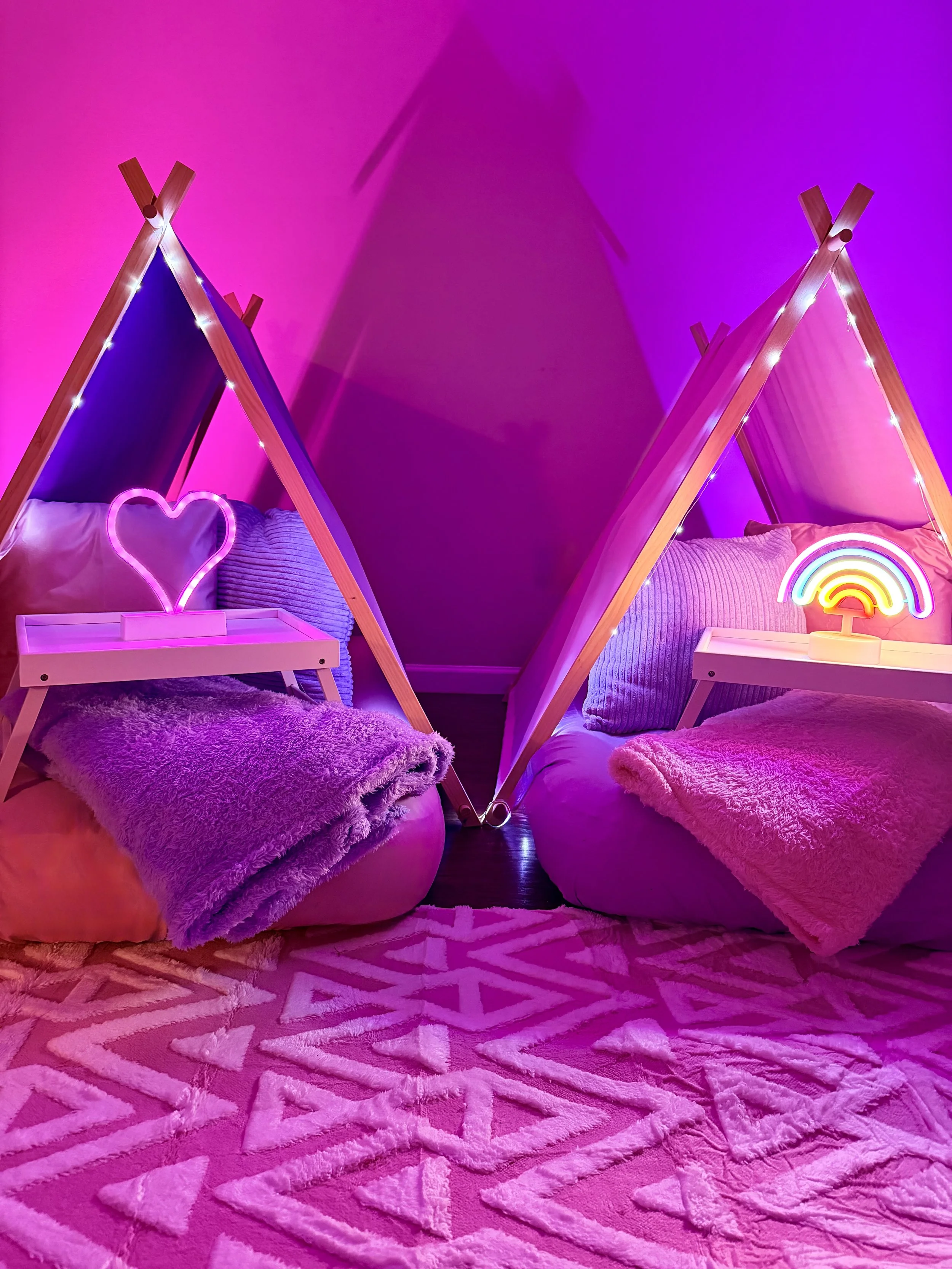 Two colorful indoor beds in a pink-themed room with neon heart and rainbow decorations.