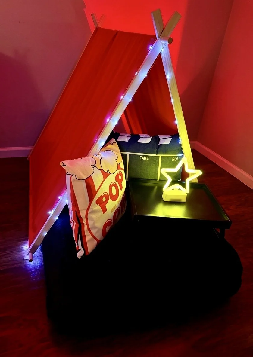 A small teepee-style tent with a red exterior and a red curtain. Inside, there is a pillow with popcorn illustration and the word "Popcorn," and a small black table with a neon star light decoration and a game board labeled "Take" and "Roll." The scene is illuminated with colorful lights.