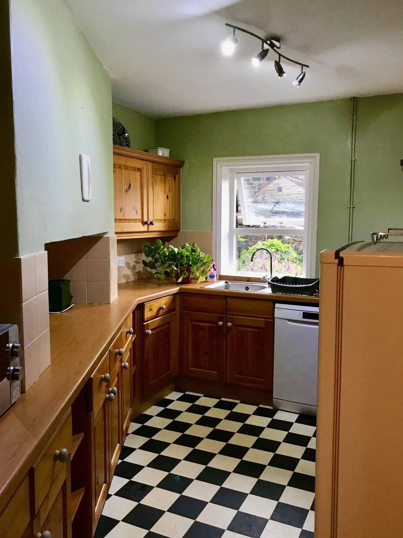 Green Gables fitted kitchen with wooden kitchen units and work surface and a black and white floor. The window looks out onto the garden.  There is a sink by the window with a dishwasher underneath.  This image also shows the toaster and the microwav