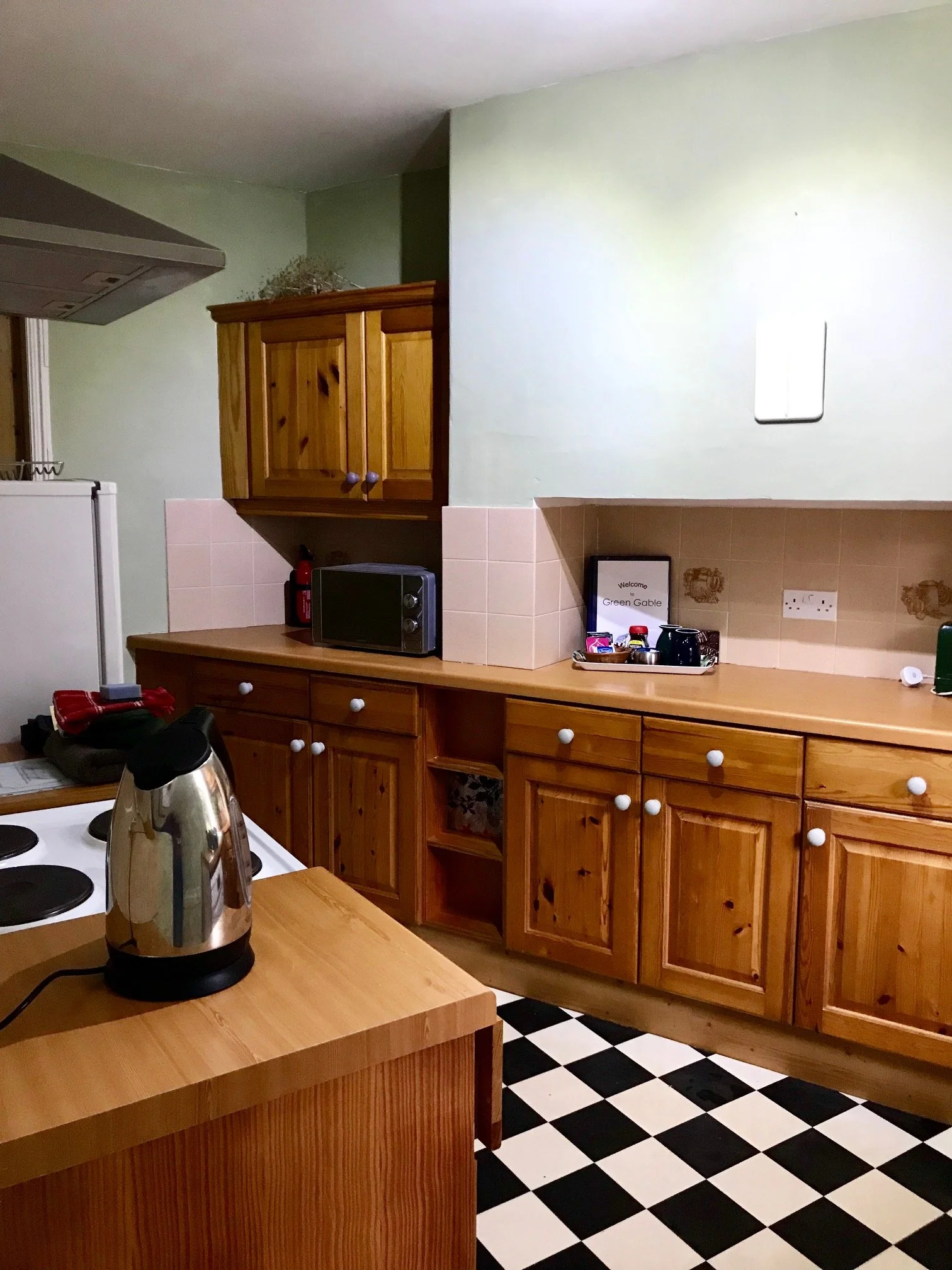 A kitchen with wooden cabinets, a black and white checkered floor, a countertop with a kettle, and a microwave. The walls are painted light green. There are various small appliances and decorations on the countertop.
