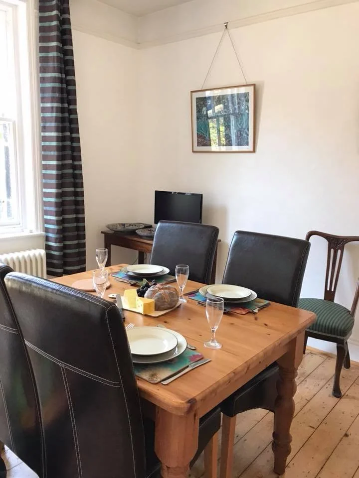 A dinning room with a wooden dining table set for four chairs with plates, wine glasses, and a centerpiece.  The room has a window with curtains, a TV, and framed prints on the wall.