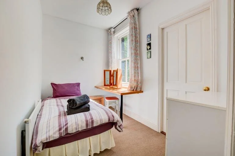 A small bedroom with a bed covered in plaid bedding, a purple pillow, and folded towels on top. There is a window with floral curtains and a small wooden desk with a mirror and a white stool. The walls are white, and the floor is carpeted.