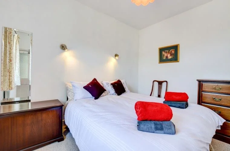 A cozy bedroom with a large bed covered in white bedding and accented with three decorative pillows. Two folded towels, one red and one gray, are neatly placed on the bed. The room features warm wooden furniture, including a dresser and a sideboard, 