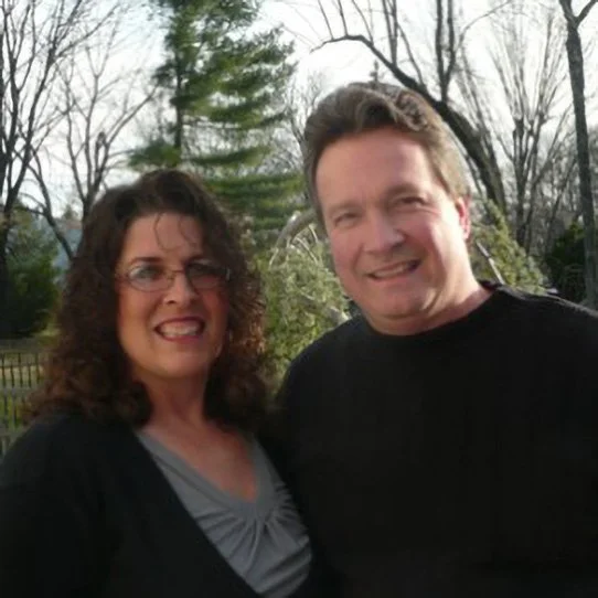 A woman and a man outdoors smiling for a photo with trees and a cloudy sky in the background.