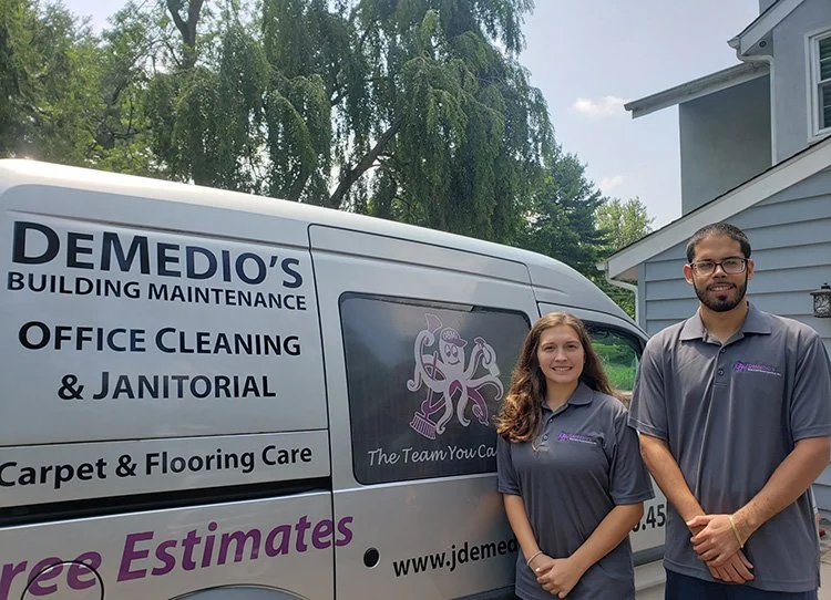 Two people standing next to a white van with business logos and text, wearing matching gray uniforms and smiling outdoors.