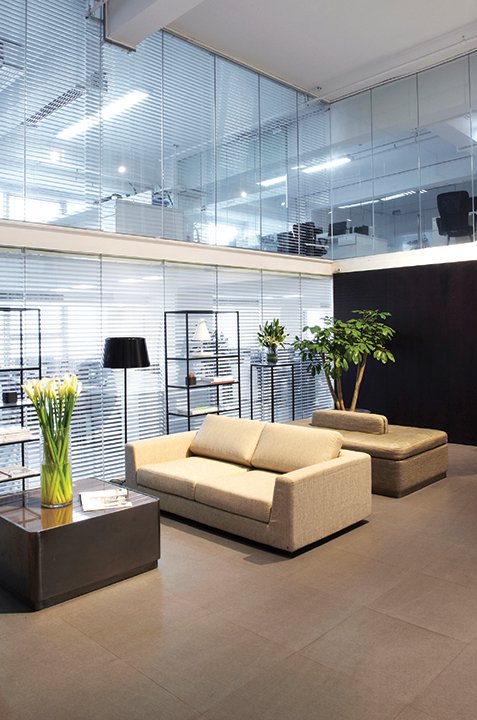 Modern office lounge with beige sofas, a black side table, a green plant, and metal shelves with lamps and decor, set against a backdrop of glass walls and blinds.