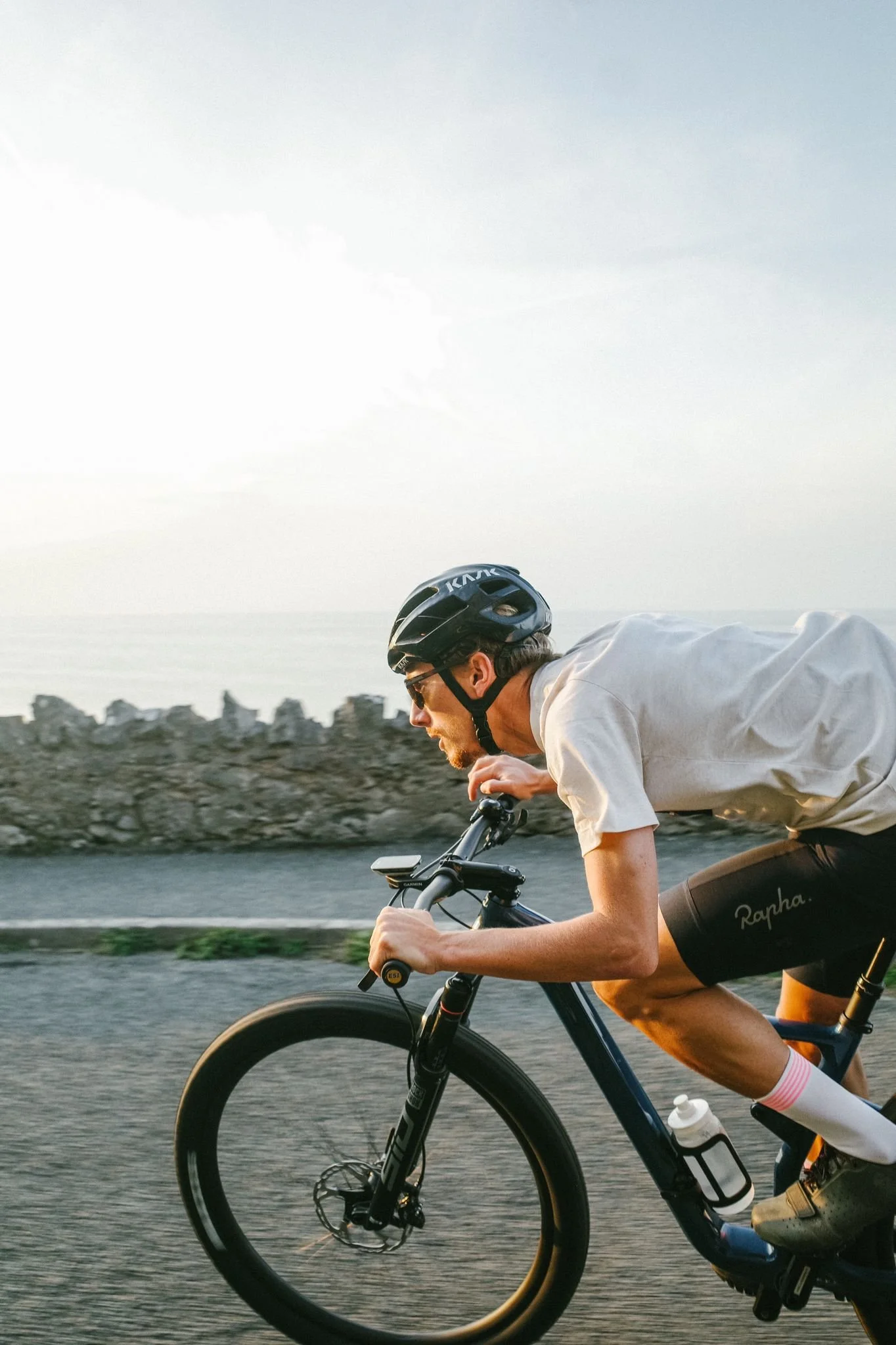 A man wearing a black helmet, sunglasses, and athletic clothes rides a mountain bike on a scenic road near water and rocks at sunset.
