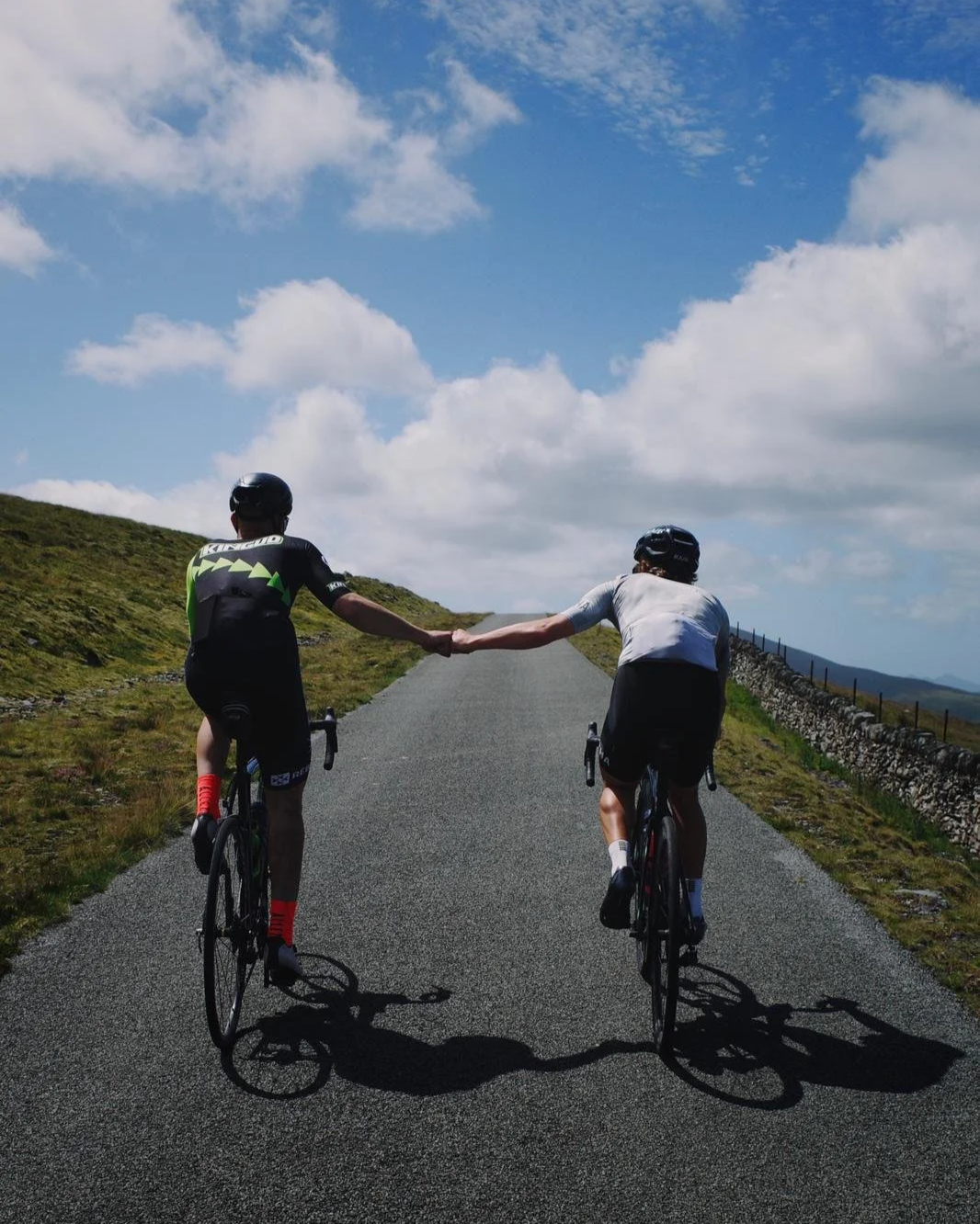 Two cyclists holding hands while riding on a paved road through a scenic landscape with grassy hills and a partly cloudy sky.