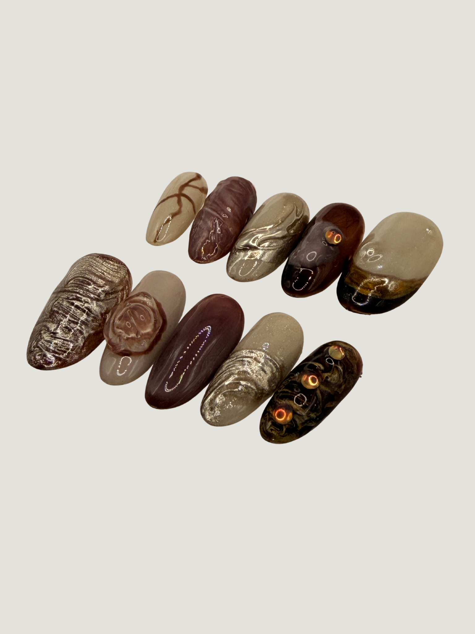 A collection of 12 decorative artificial nails with marbled and textured designs in shades of beige, brown, black, and gold, arranged on a light background.