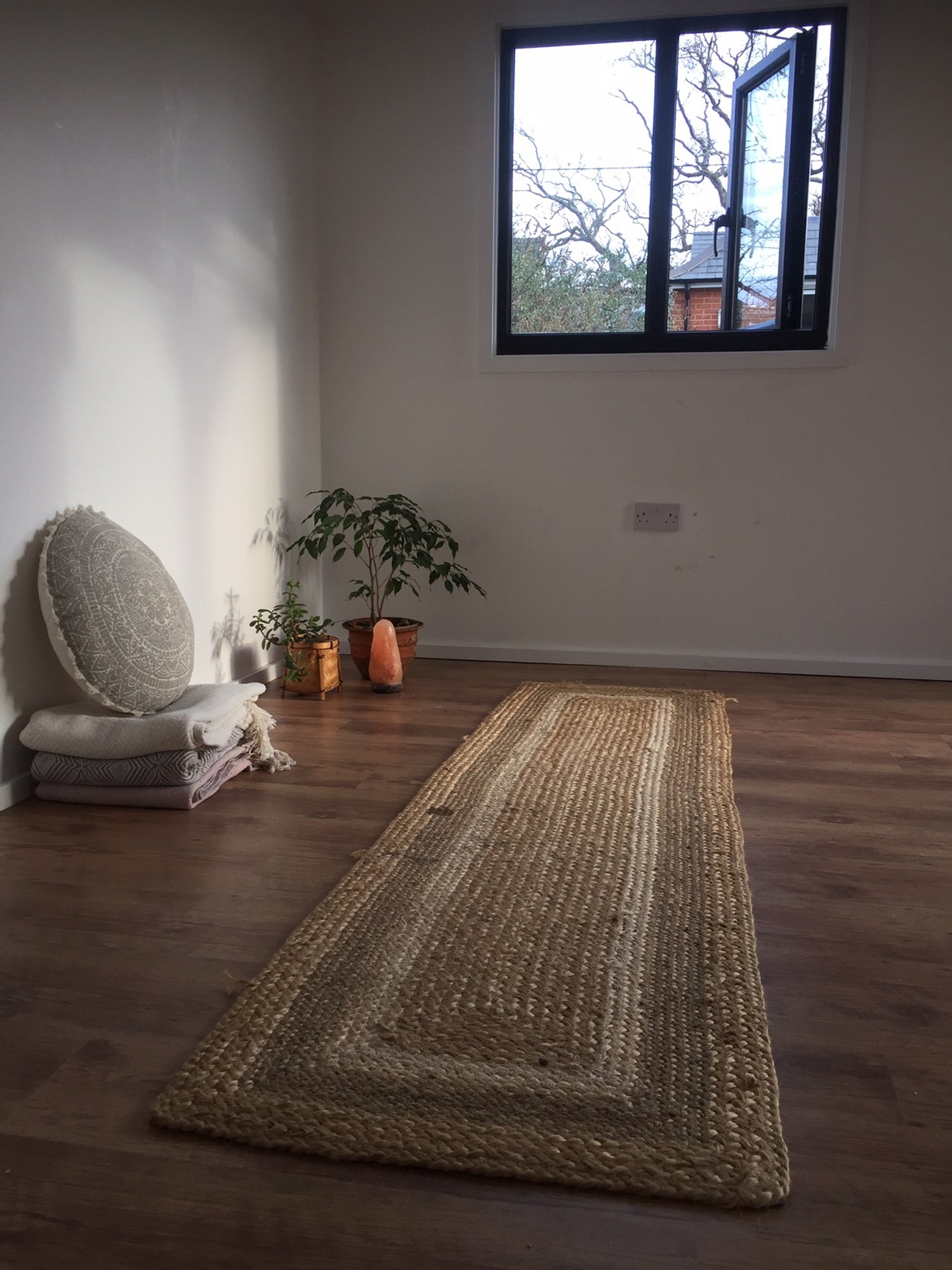 A minimalistic room with hardwood flooring, a window with an open black frame, a potted plant near the wall, a folded blanket with a round decorative cushion, a salt lamp, and a beige woven rug running along the floor.