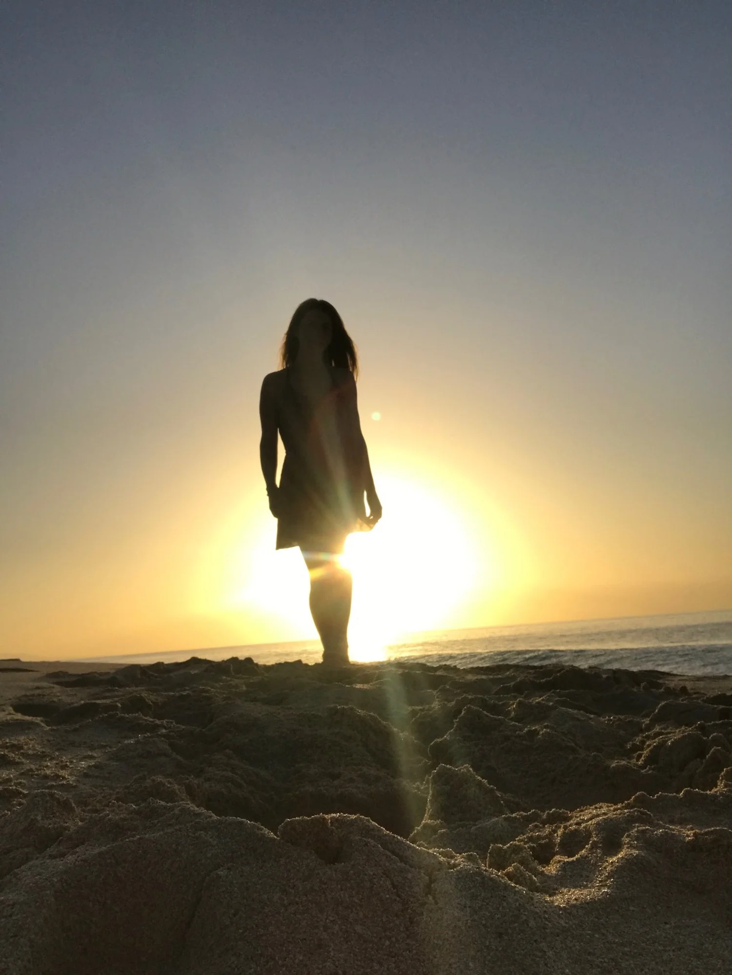 Silhouette of a woman standing on a sandy beach at sunset or sunrise with the sun behind her, creating a glowing background and lens flare.