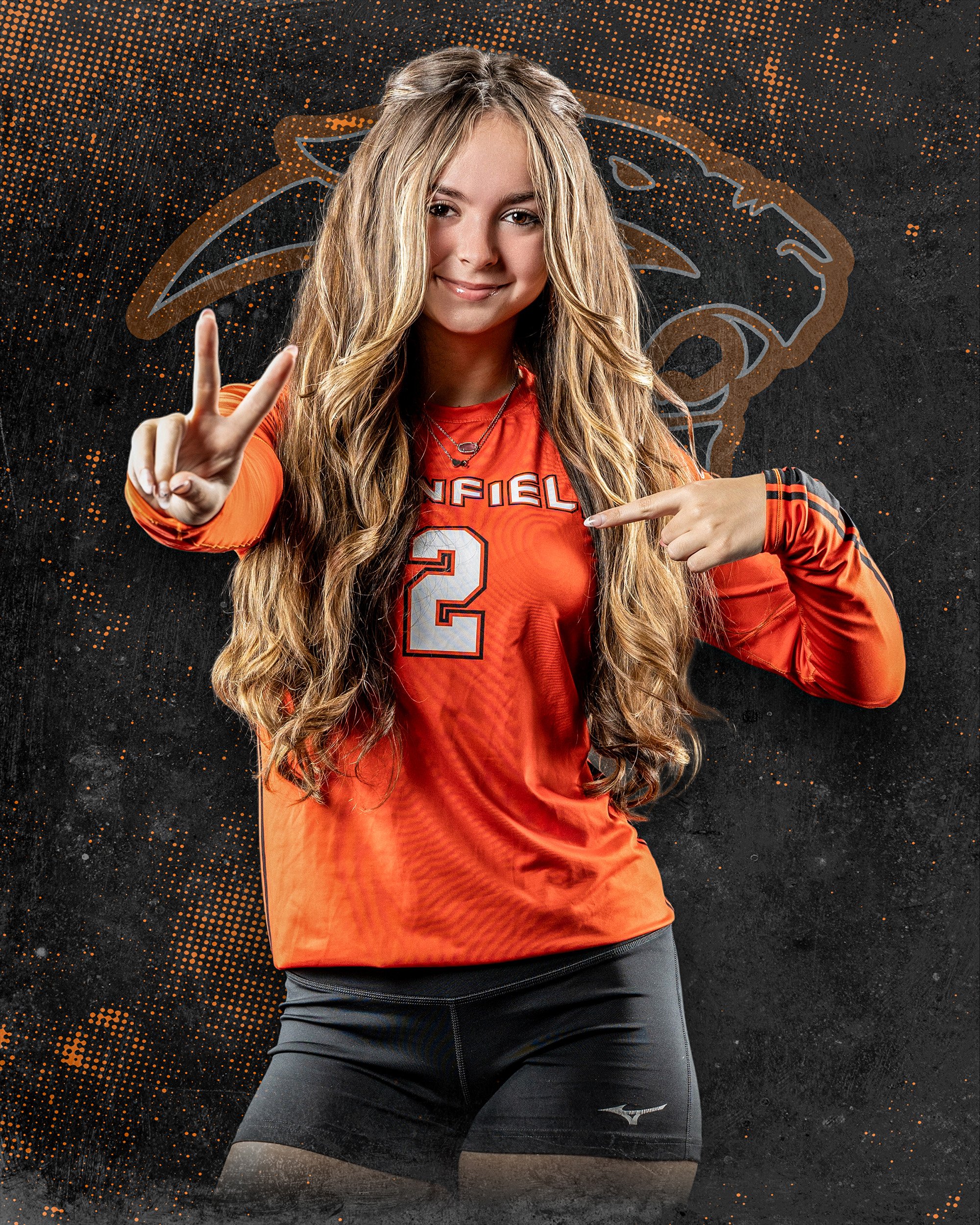 A young woman in a red sports jersey with the number 2 and the name 'DANFIELD' on it, smiling and making a peace sign with her right hand, pointing at her jersey with her left hand. She has long, wavy blonde hair and is standing against a dark backgr