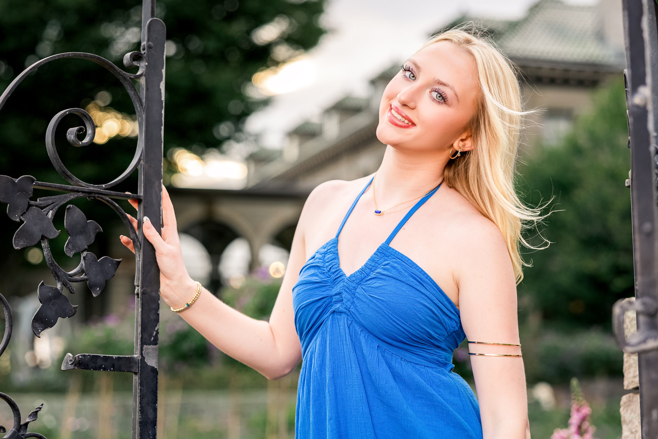 A young woman with blonde hair and blue eyes smiling while standing outdoors, holding a decorative black metal gate, wearing a blue dress and jewelry.