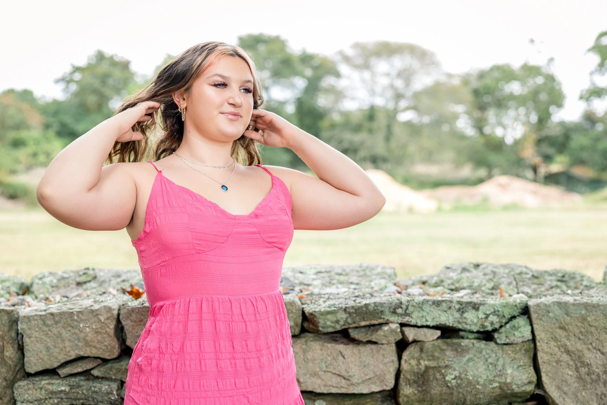 Young woman outdoors in a pink dress, adjusting her hair, standing in front of a stone wall with trees in the background.