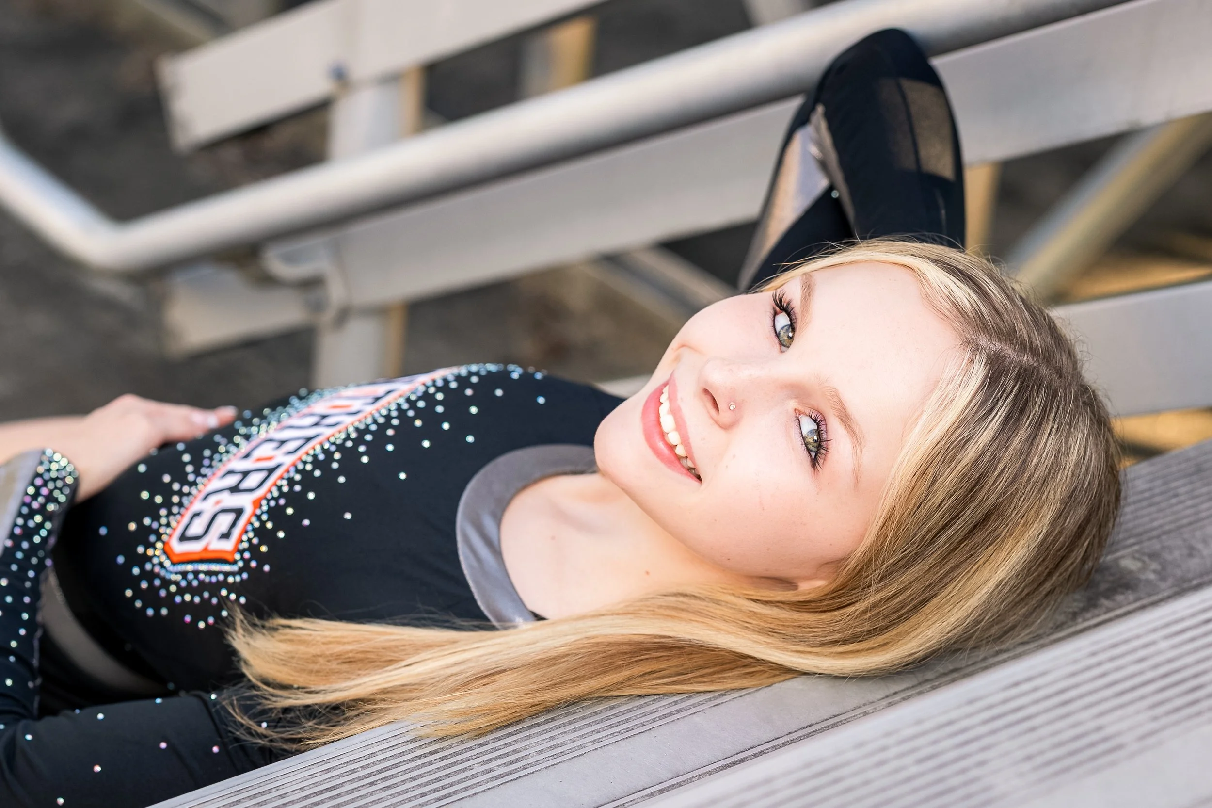 A young woman with long blonde hair and blue eyes lying on a wooden bench, smiling at the camera, wearing a black shirt with rhinestones and a spicy denim logo.