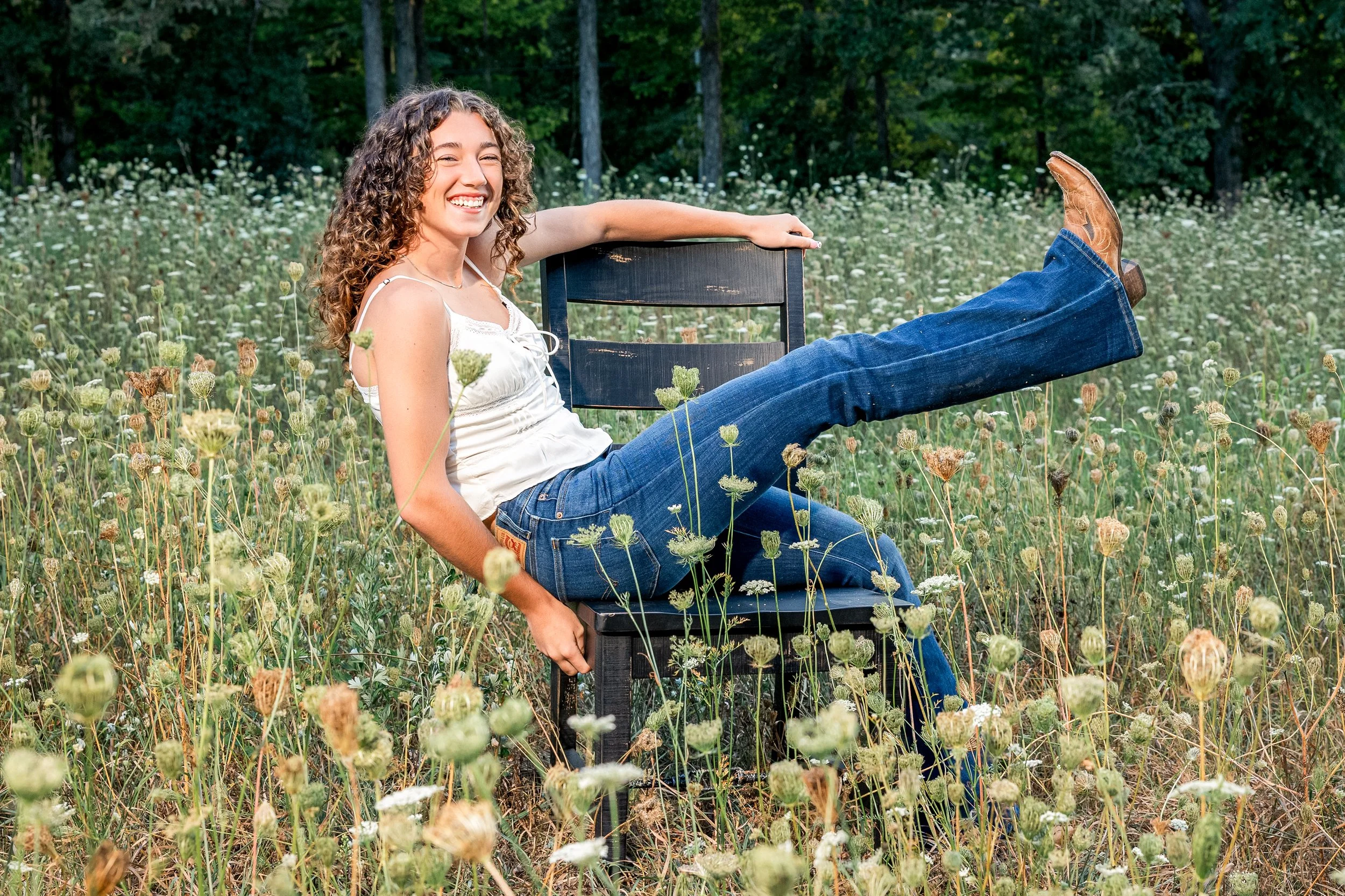 A young woman with curly hair sitting on a black chair in a green field of wildflowers, smiling and posing with one leg extended.