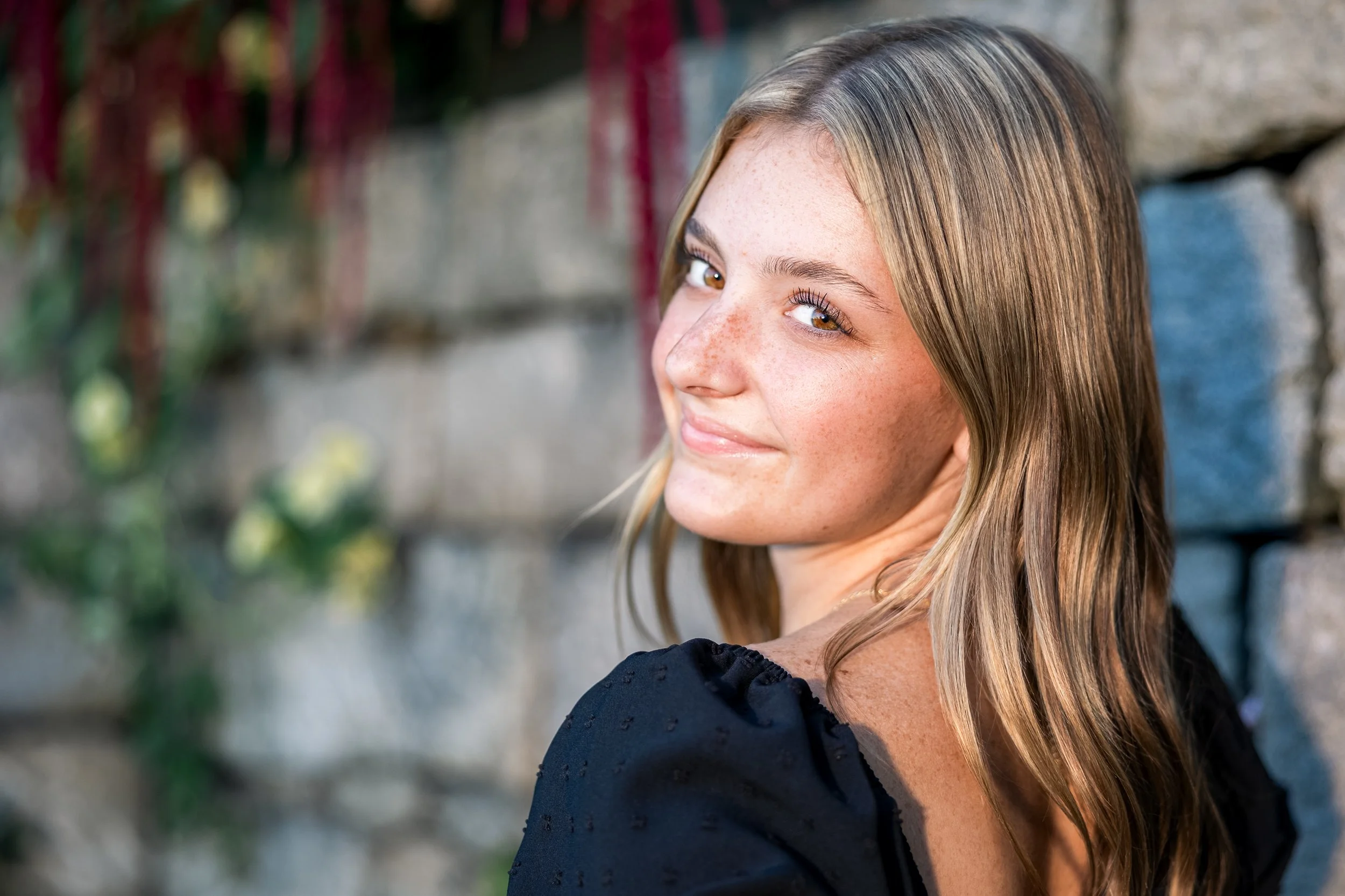 A young woman with long blond hair and freckles, smiling at the camera, against a blurred stone wall background with greenery.