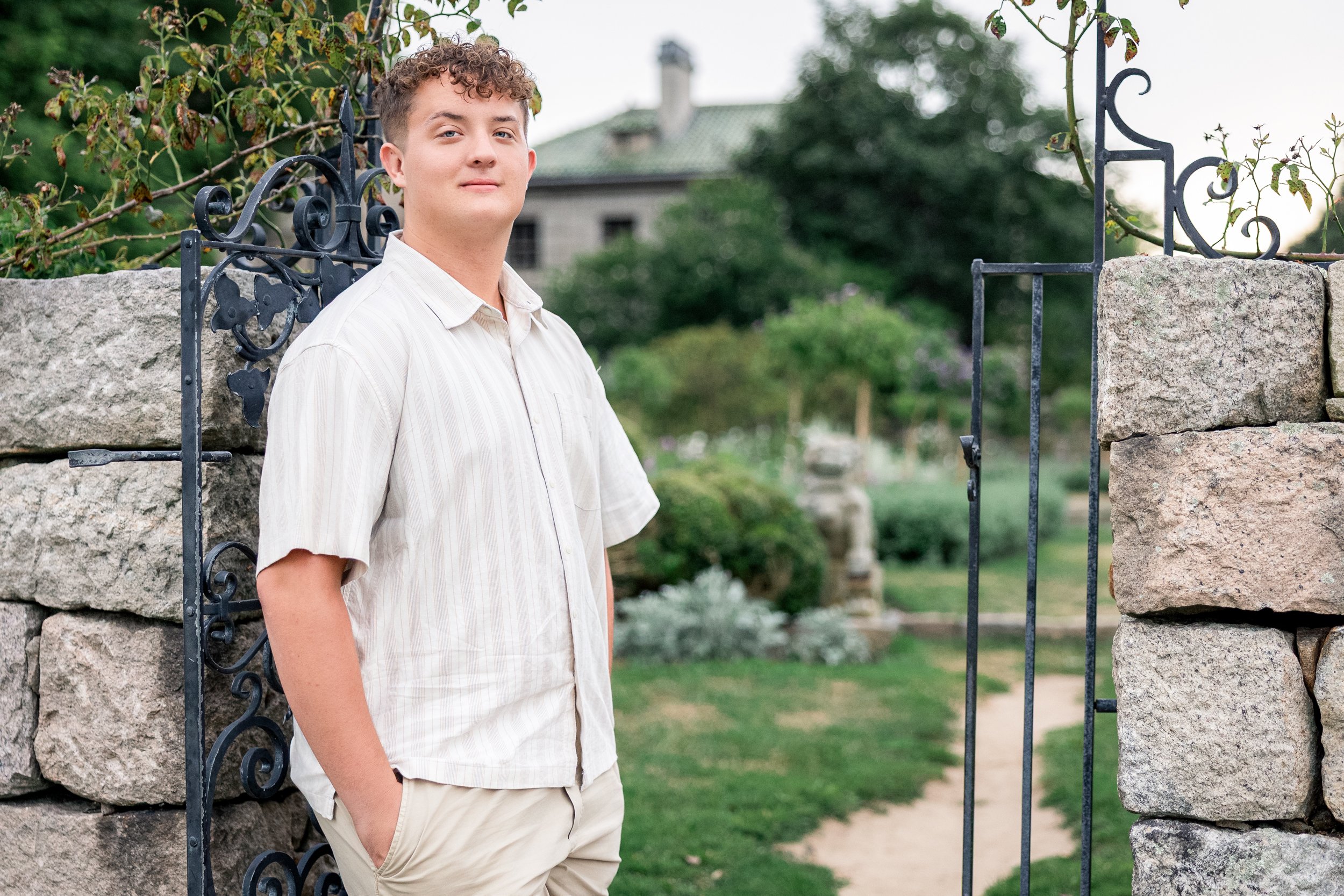 A young man with curly hair wearing a beige striped shirt and khaki pants standing at an open wrought iron gate in front of a stone wall, with a garden and a house in the background.
