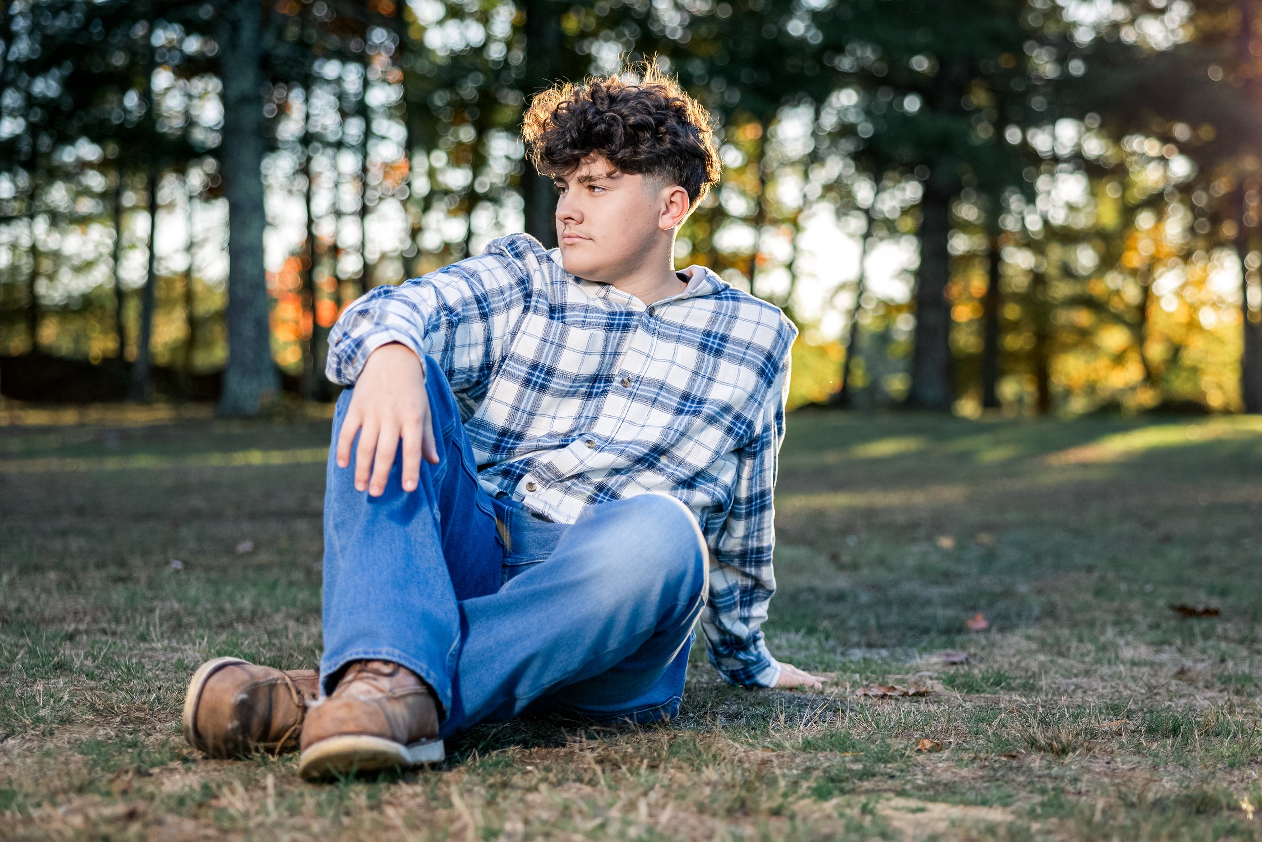 Young man with curly hair sitting on the grass in a park during sunset, wearing a plaid shirt, blue jeans, and brown boots.