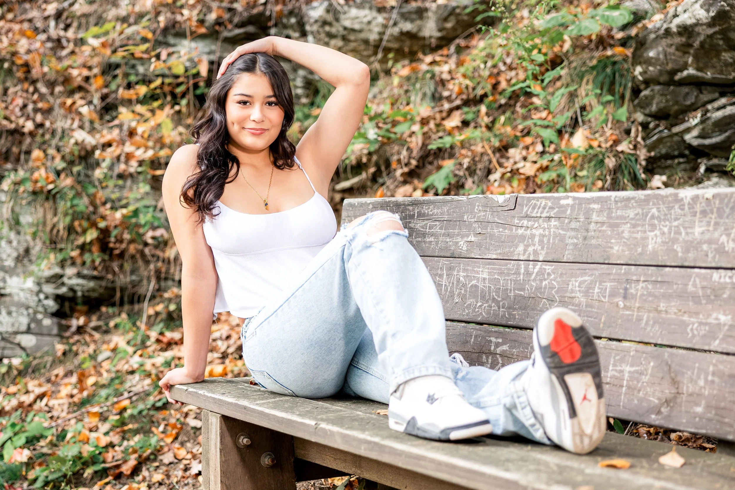 Young woman with dark wavy hair sitting on a wooden park bench surrounded by fallen autumn leaves, wearing a white tank top, light blue jeans, and white sneakers, smiling and looking at the camera.