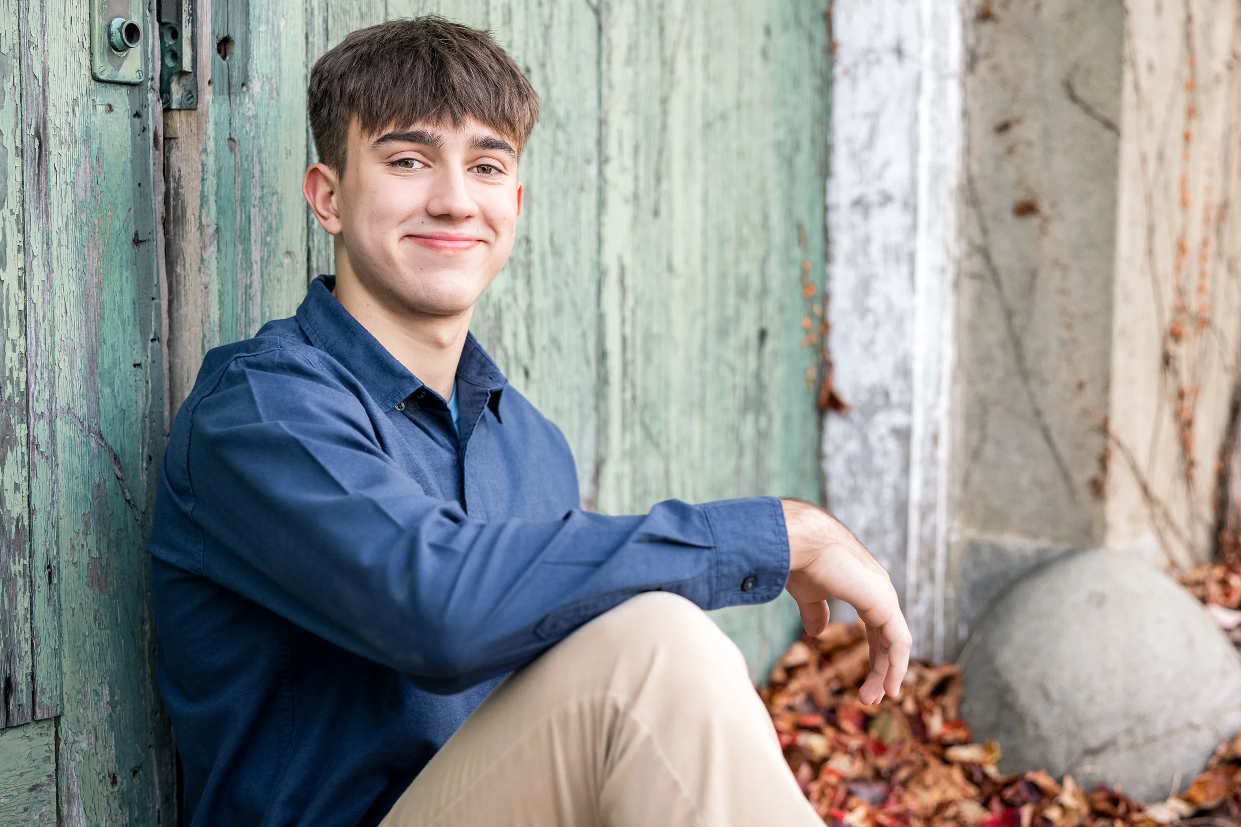 A young man sitting outdoors next to a weathered wooden wall with green and white paint, surrounded by fallen autumn leaves and a large round stone.