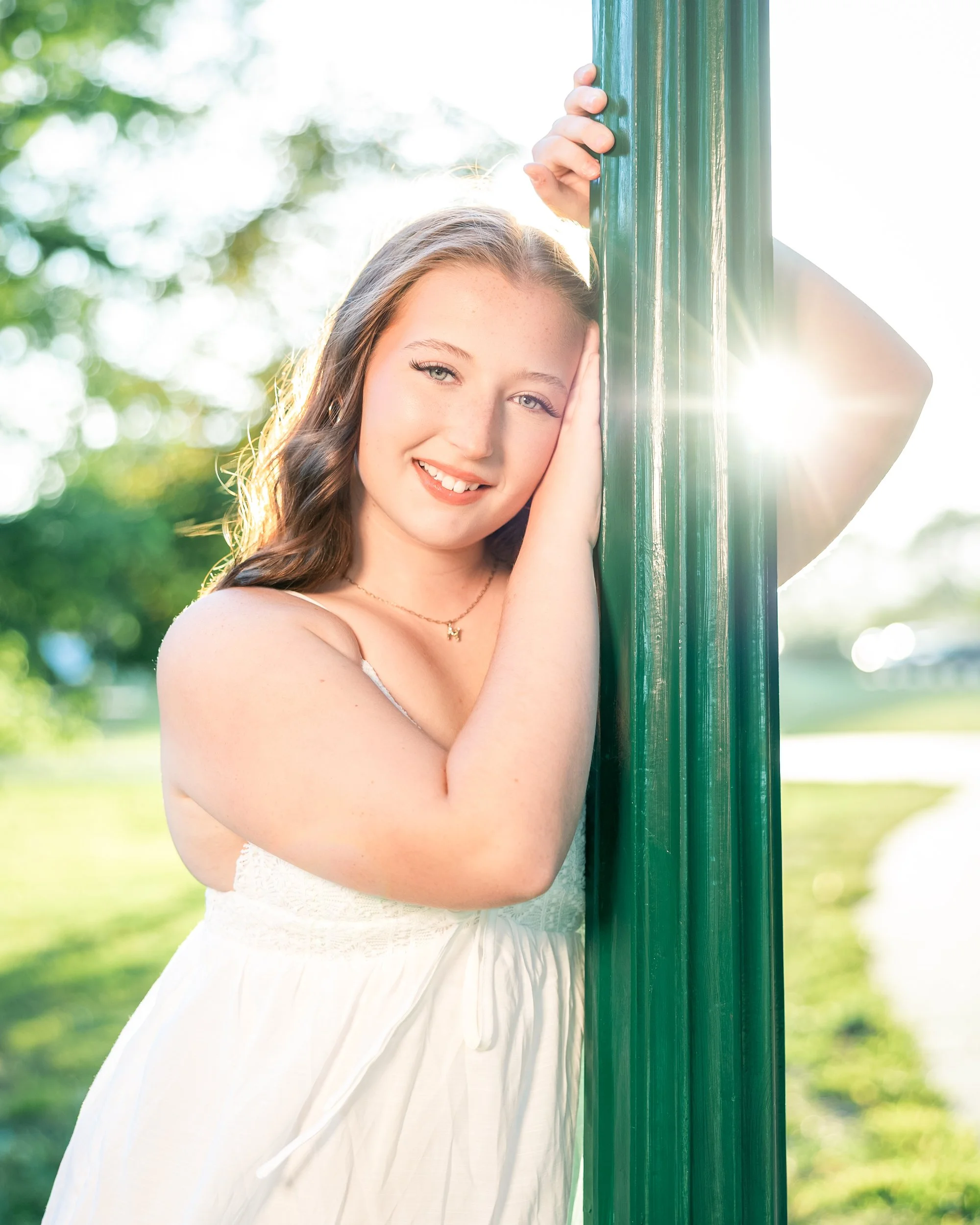 A young woman with long brown hair and blue eyes smiling, wearing a white dress, standing outdoors in sunlight, holding a green pole with one hand and resting her face on the other, in a park with trees and grass in the background.