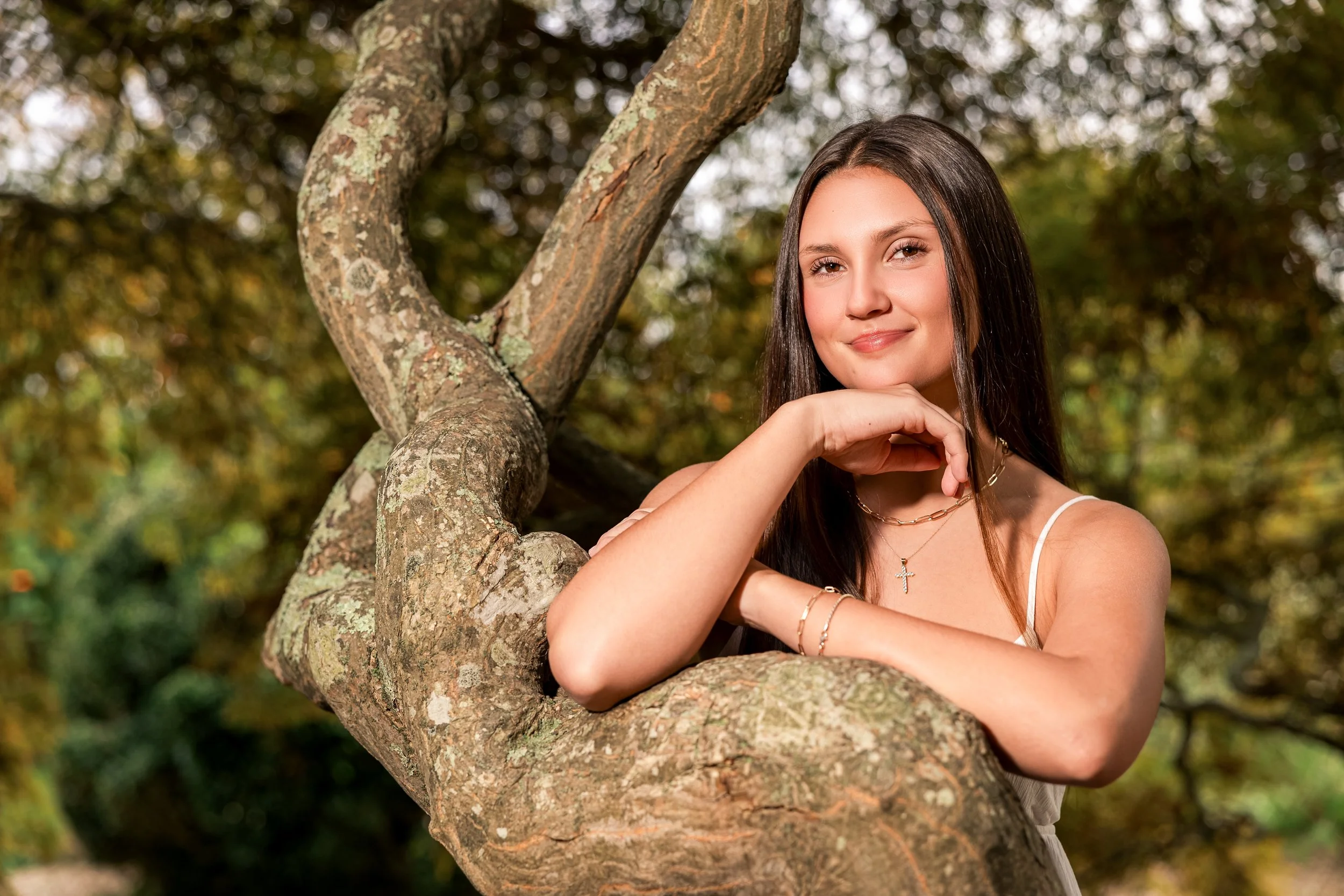 A young woman with long dark hair and light skin leaning on a tree branch outdoors, smiling softly, wearing a white sleeveless dress, jewelry including a cross necklace and bracelets, with autumn foliage in the background.