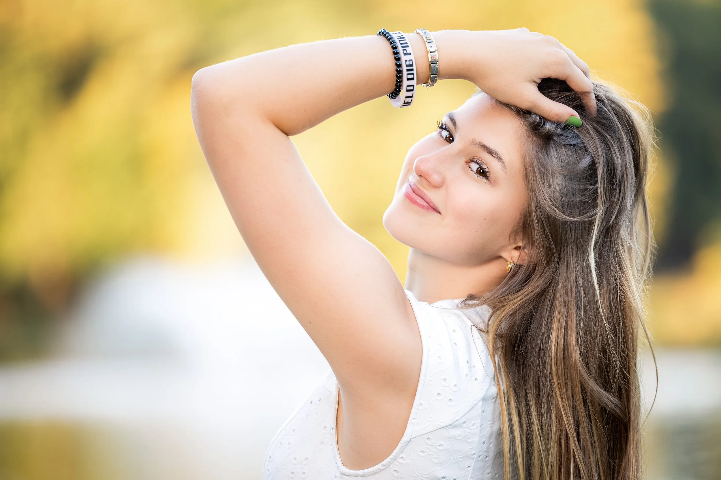 Young woman with long brown hair smiling outside, wearing a white top and bracelets, with a hand on her head.