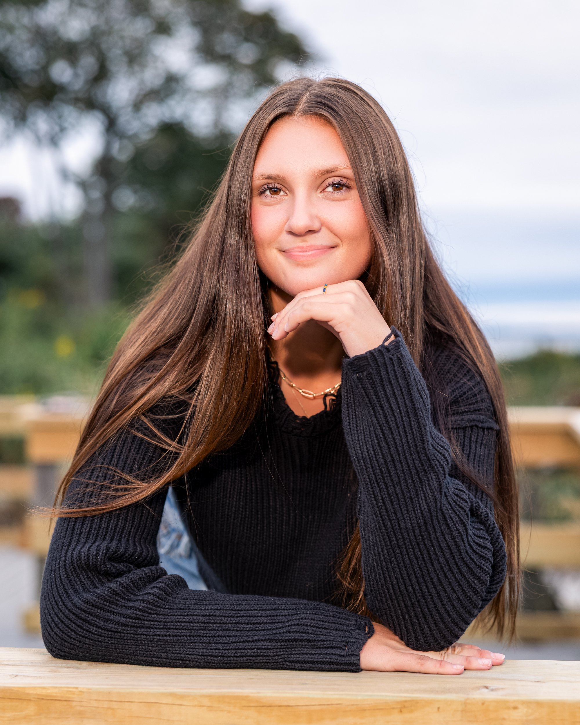A young woman with long brown hair sitting outdoors at a wooden table, resting her chin on her hand, smiling at the camera. She wears a black sweater and layered necklaces, with a blurred natural background.