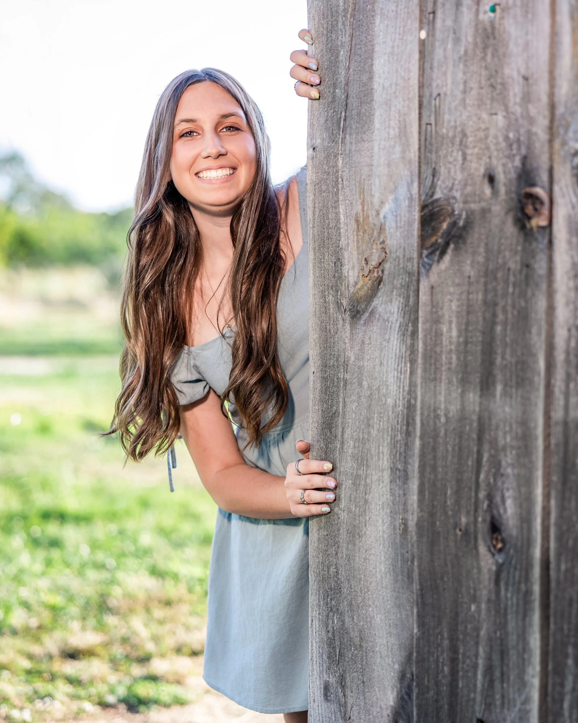 A woman with long brown hair smiling and peeking from behind a wooden fence outdoors on a sunny day.